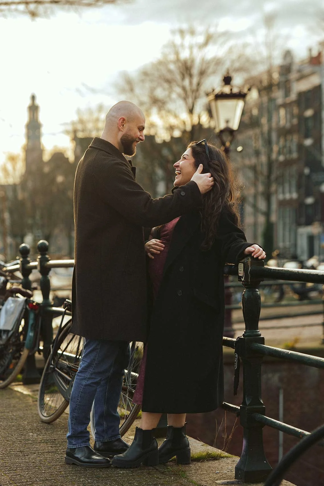 A couple embracing and smiling at each other on a bridge over a canal, with buildings in the background. The woman is pregnant