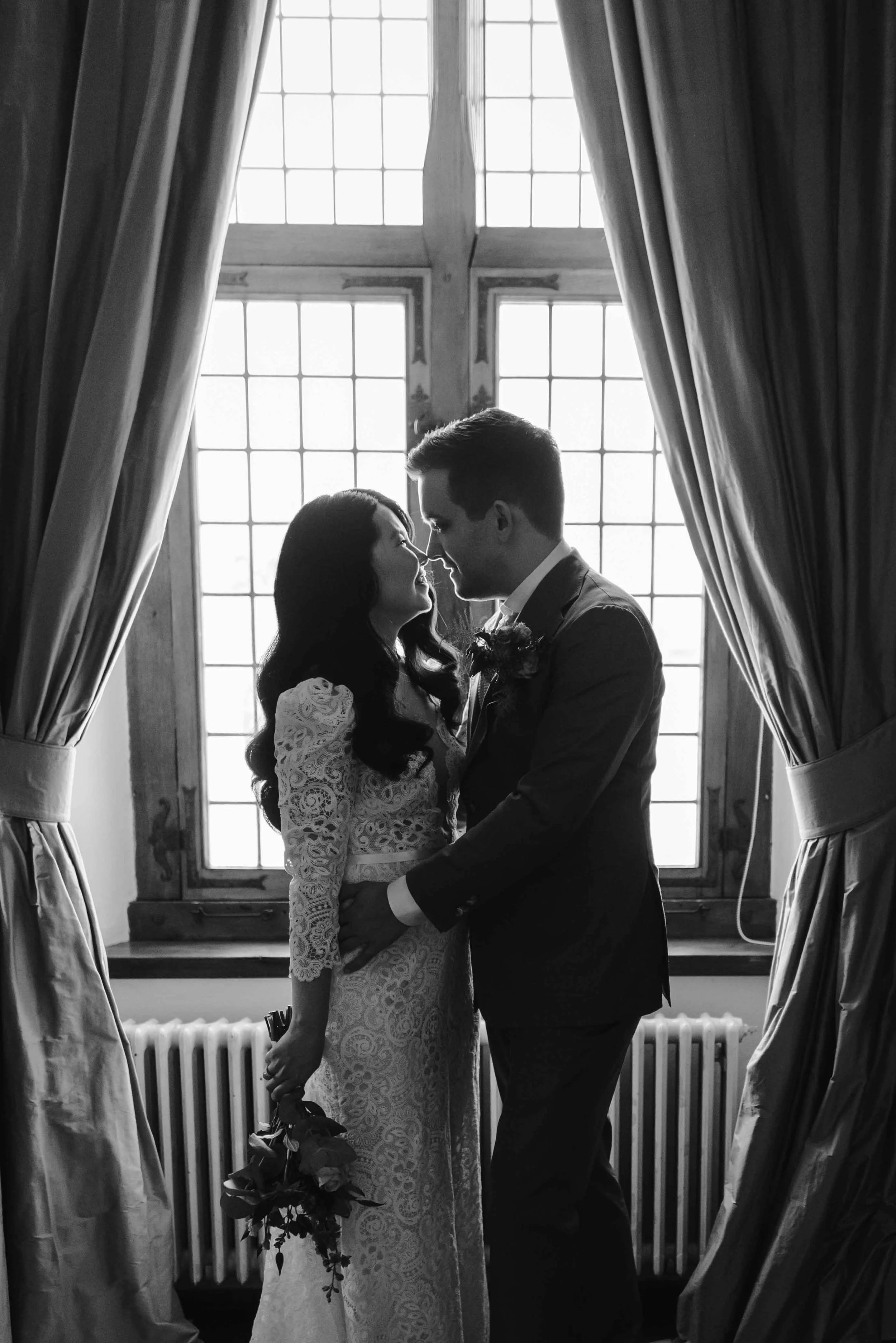 A black and white photo of a bride in a lace dress and a groom in a suit looking at each other intimately, silhouetted against a bright window with large curtains on either side.