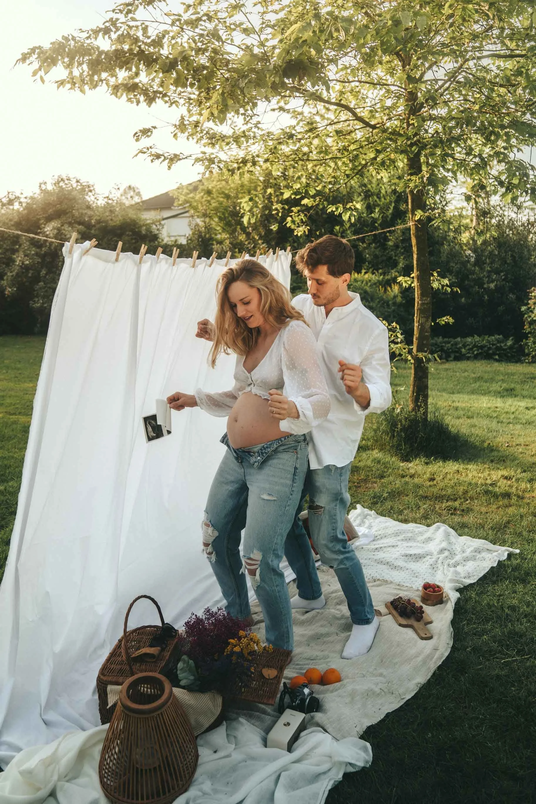 A pregnant woman and a man standing outdoors in front of a white sheet hanging on a line. The woman is holding an ultrasound photo, and the man has his arm around her.