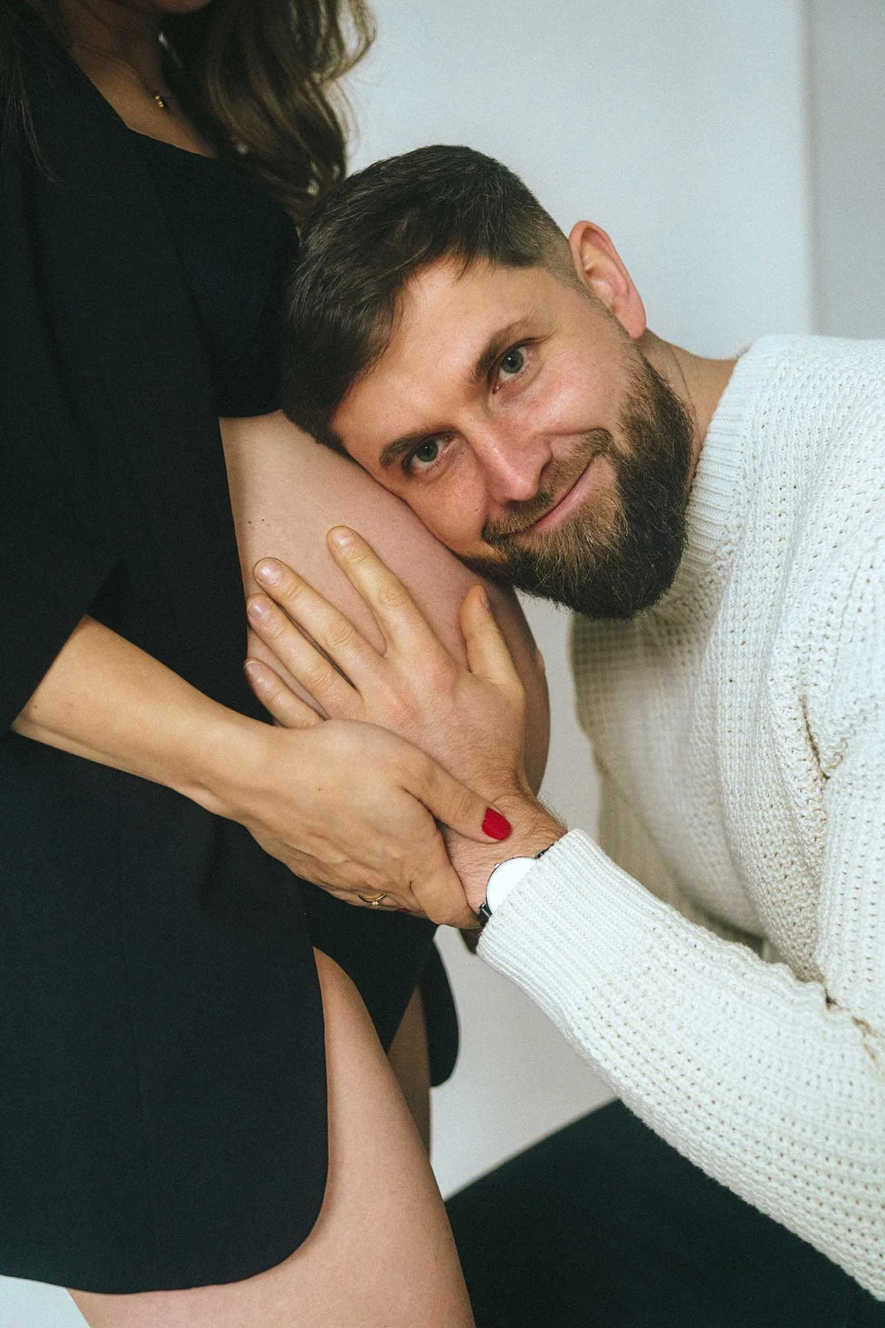 A man with a beard is smiling as he rests his head against a pregnant woman's belly, holding it gently with his hand. The woman is wearing a dark dress