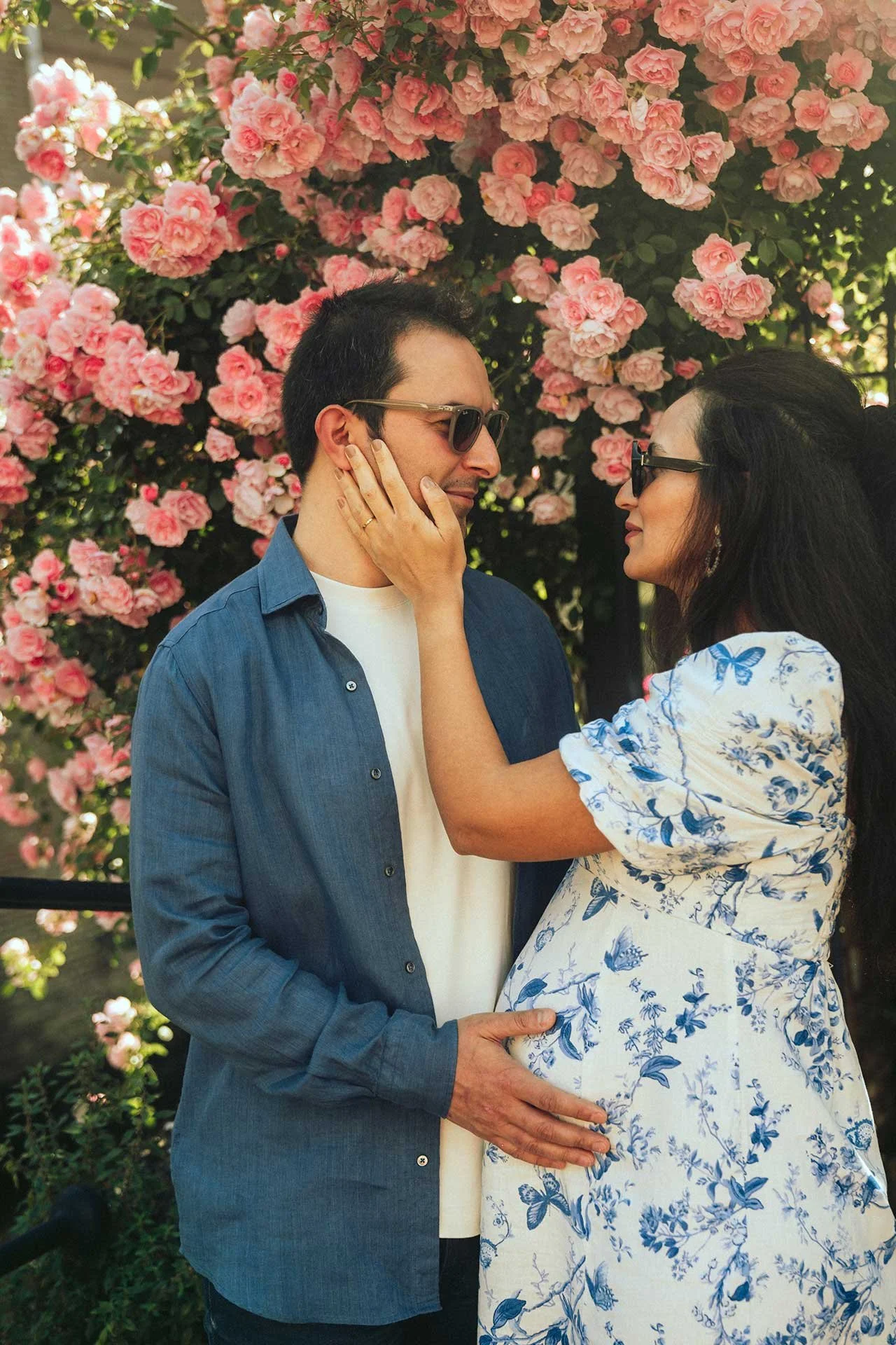 A pregnant woman in a floral dress caresses a man's face as they stand close together in front of a bush with pink flowers, both smiling at each other
