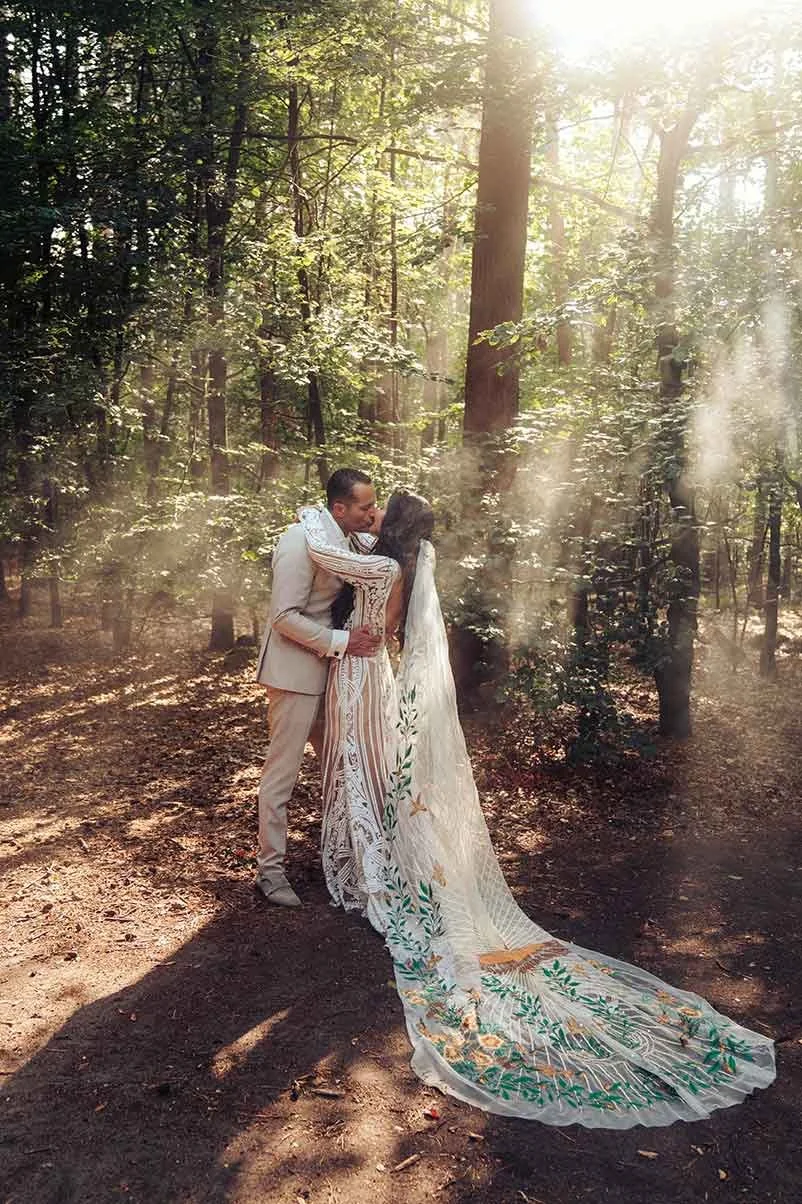 A newlywed couple embraces and kisses in a sun-dappled forest, with light rays shining through the trees. The bride wears a white gown with an elaborately embroidered long train.