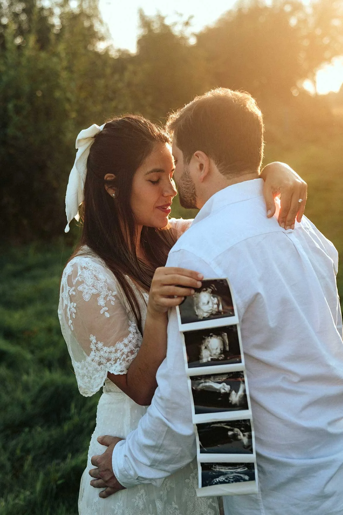 A couple embracing outdoors in warm sunlight. The woman is holding a strip of ultrasound photos of their baby