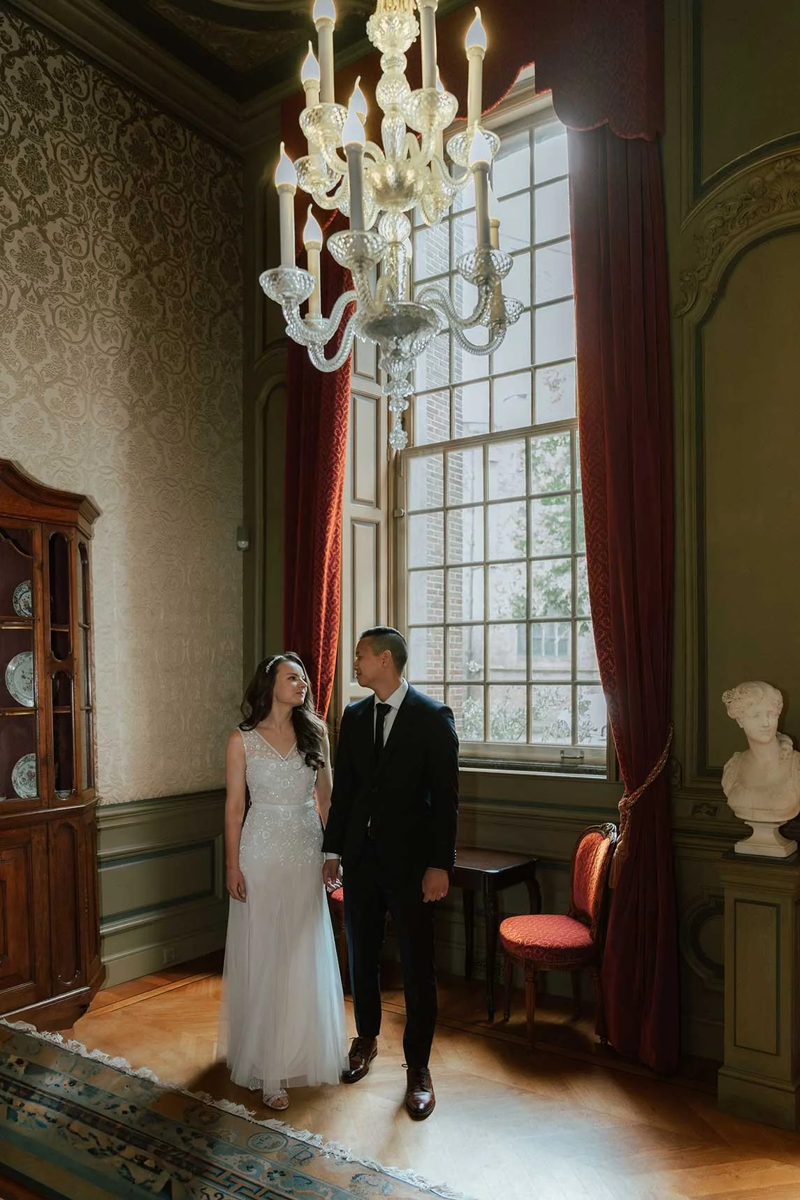 A tender glance between a bride and groom in an opulent room with a large window and chandelier, capturing the elegant atmosphere of a historic wedding location in the Netherlands