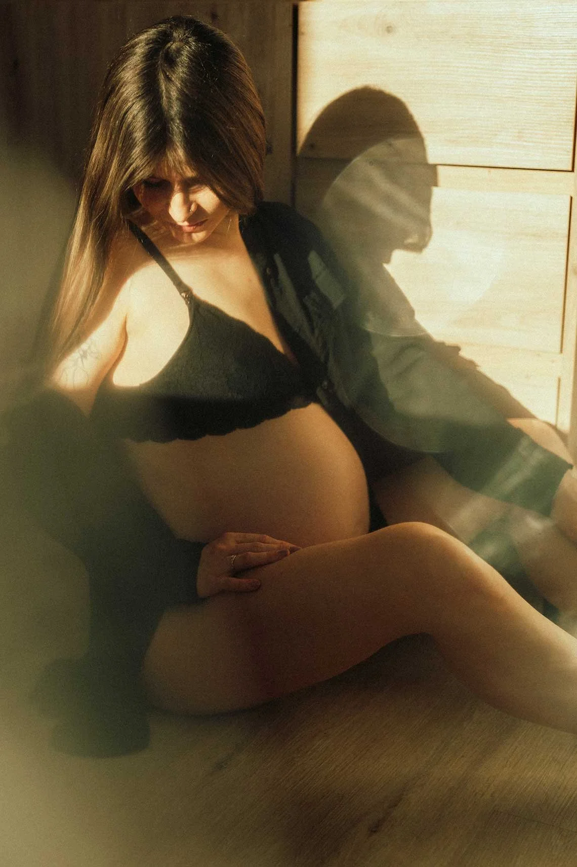 A pregnant woman sitting on the floor, looking down at her baby bump and cradling it with her hands. She is wearing a black bra and an open black shirt, with sunlight casting shadows