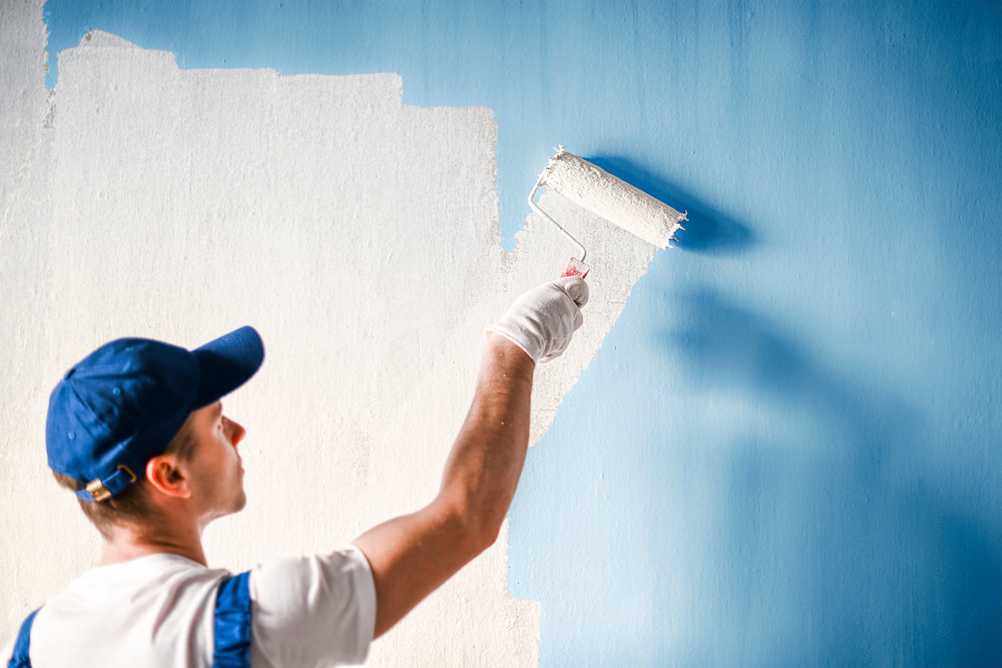 Person painting a blue wall white with a roller brush.