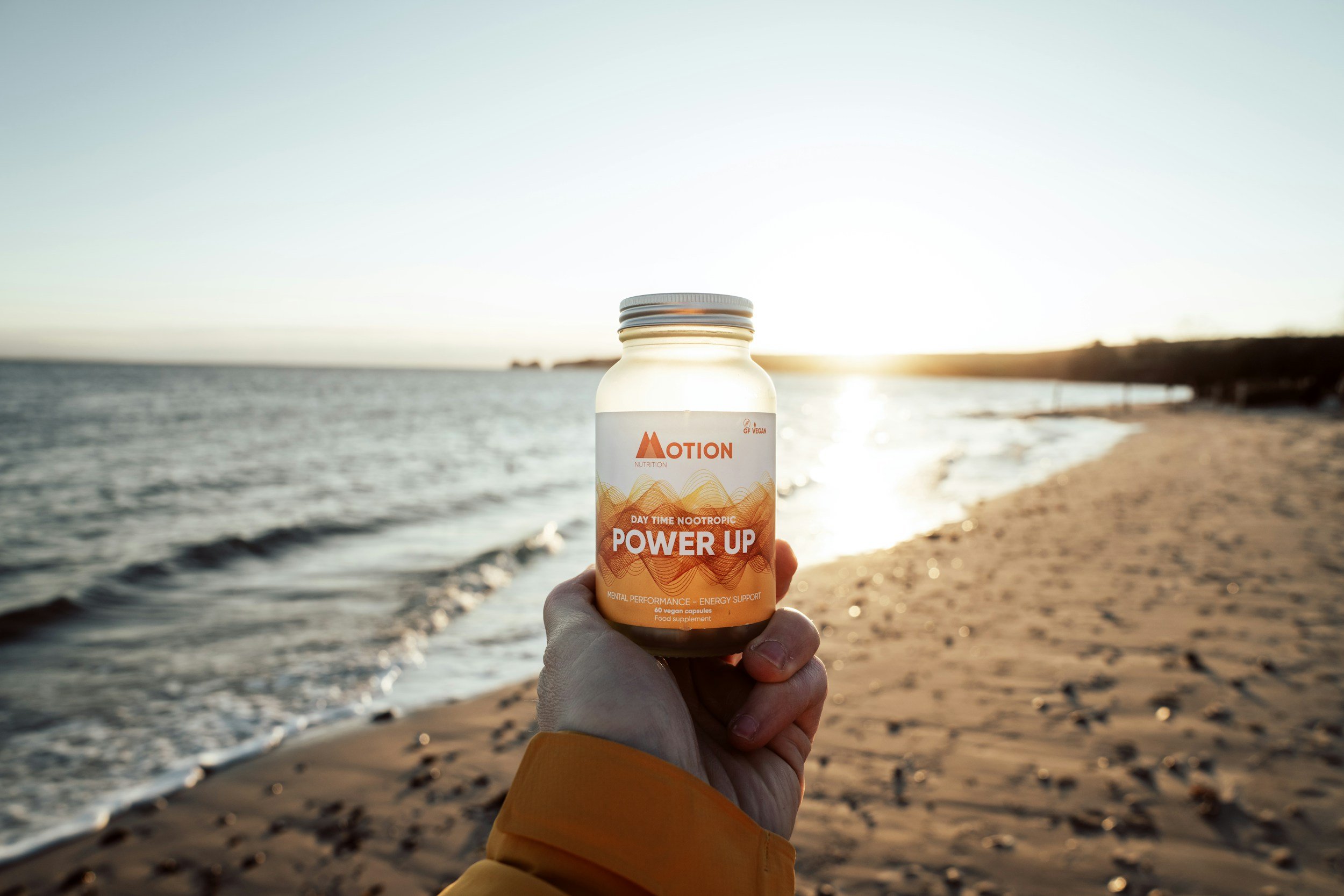 A hand holding a bottle labeled 'Power Up' on a beach at sunset with ocean waves and sandy shore in the background.