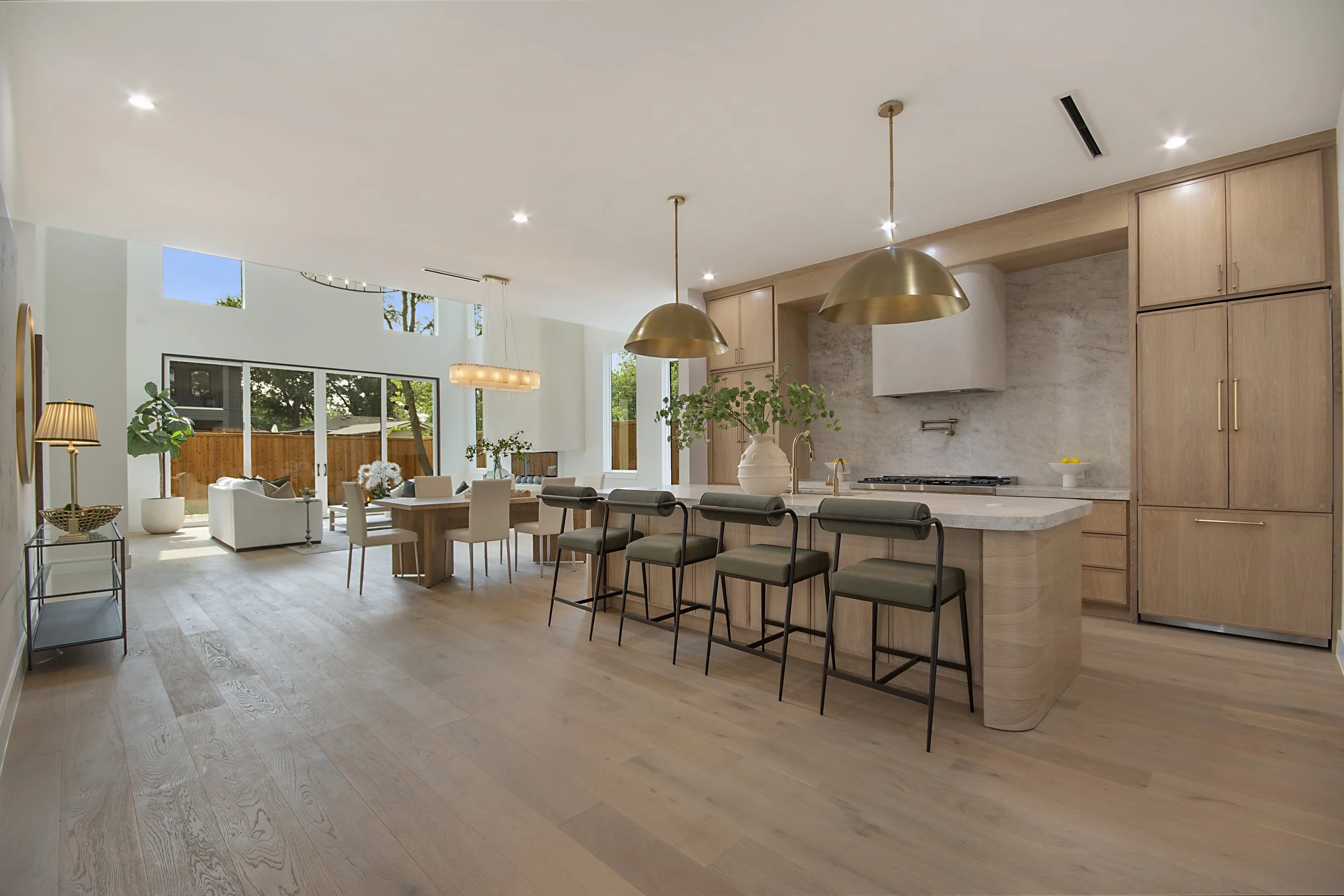 Open-concept kitchen and living room with wooden flooring, modern lighting, and large windows showing outdoor trees.