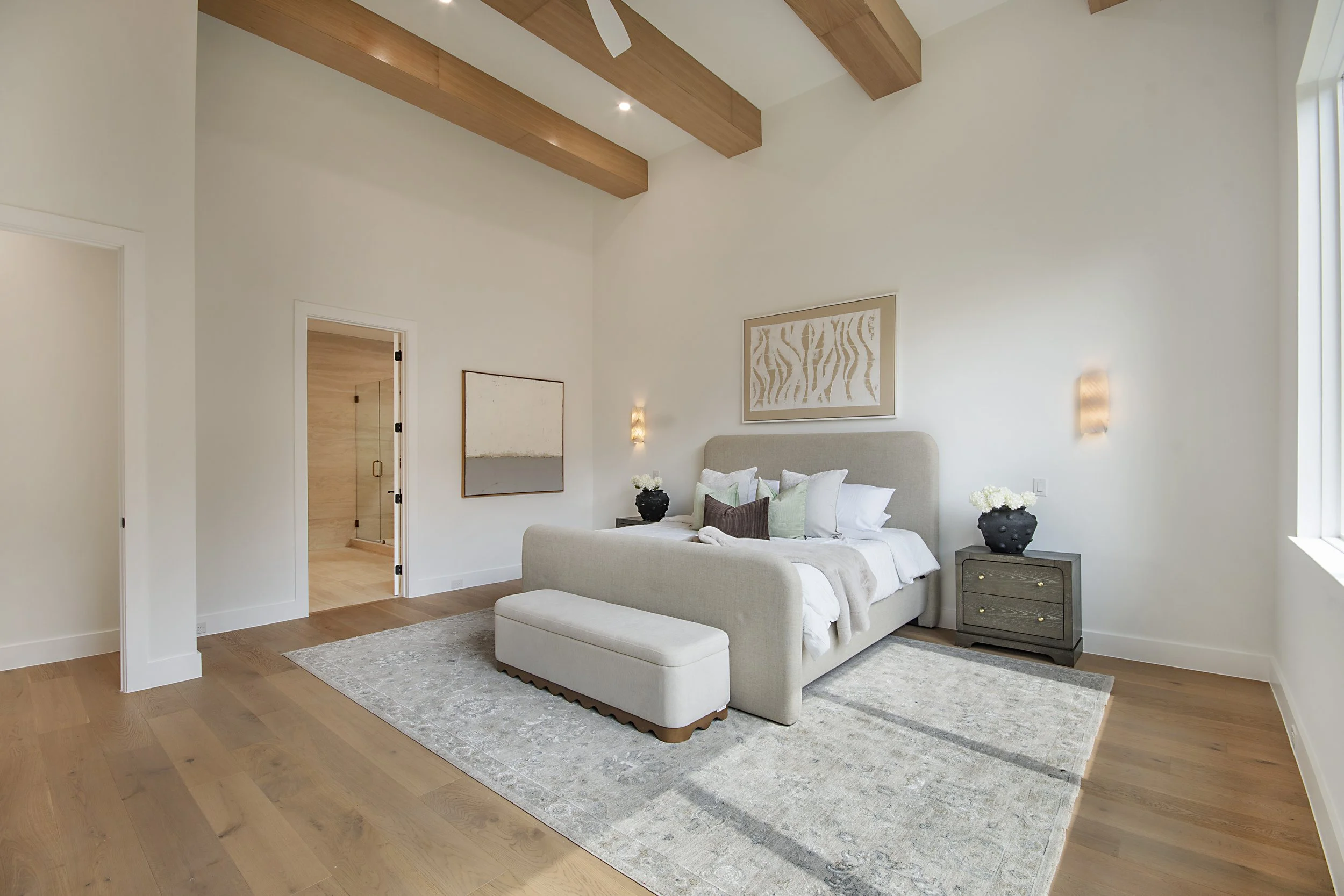 A modern bedroom with a beige upholstered bed, decorative pillows, a light-colored lamp on a wooden nightstand, artwork on the wall, and hardwood floors.