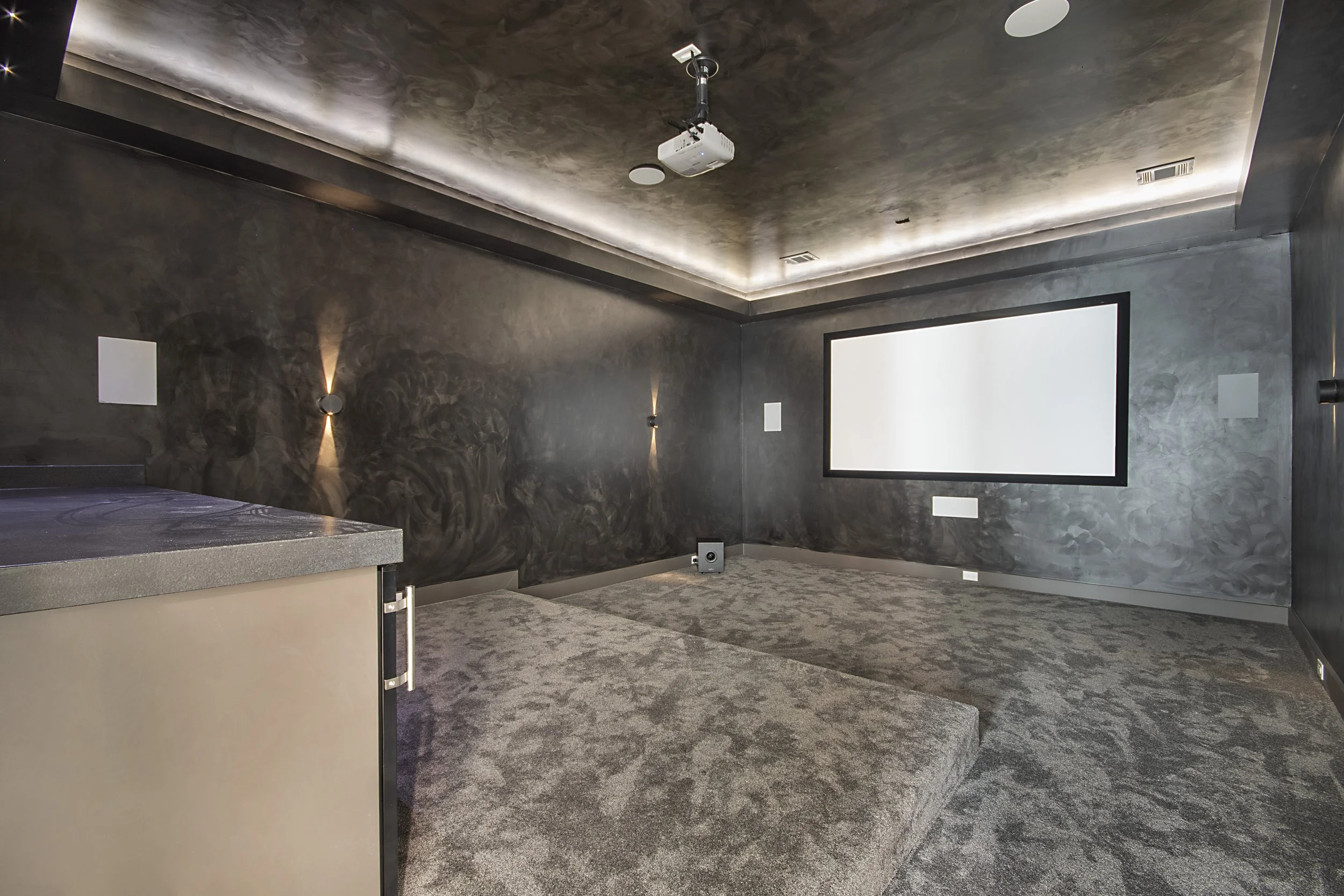 Room with dark grey textured walls and ceiling, carpeted floor, wall sconces, a large blank screen or whiteboard, and a ceiling-mounted projector.