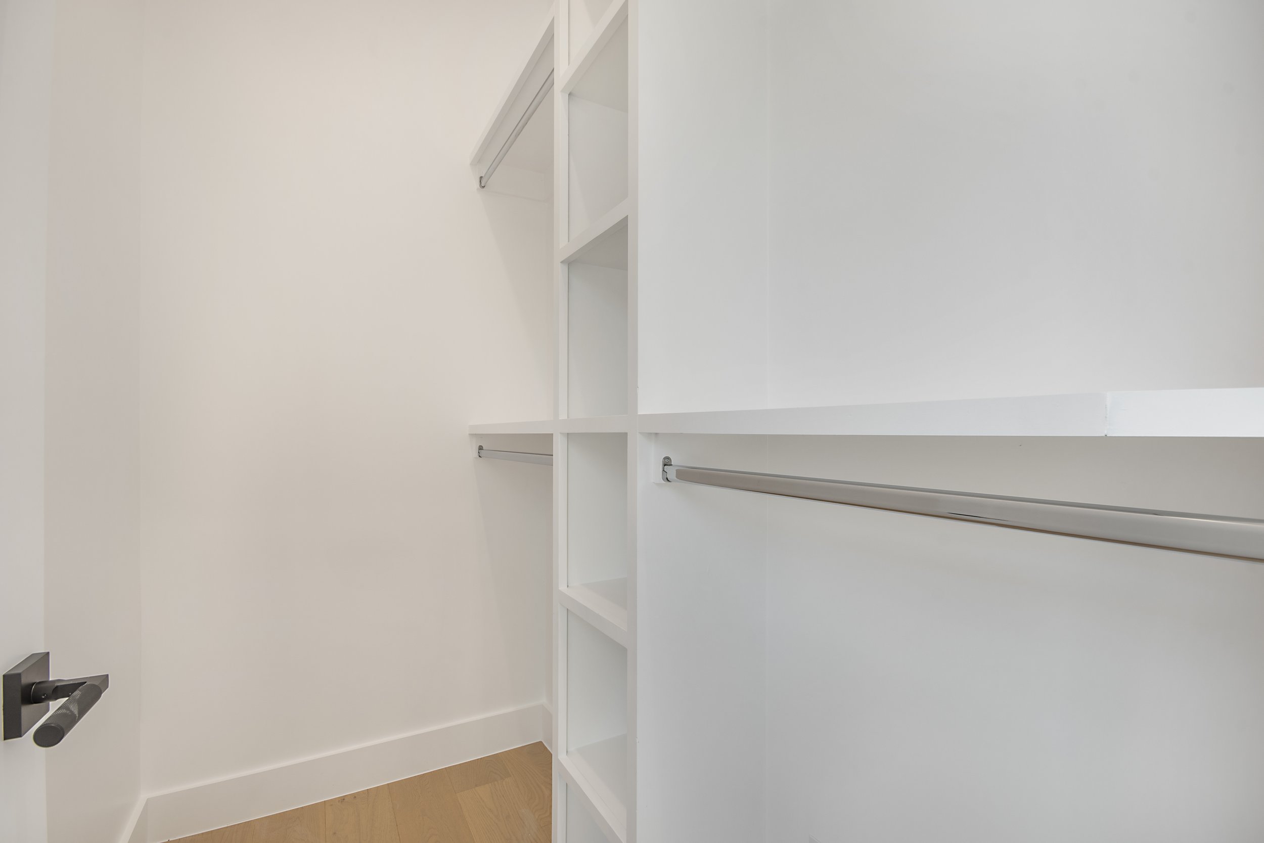 Empty white closet with built-in shelves and metal closet rods, open door showing a hardwood floor.