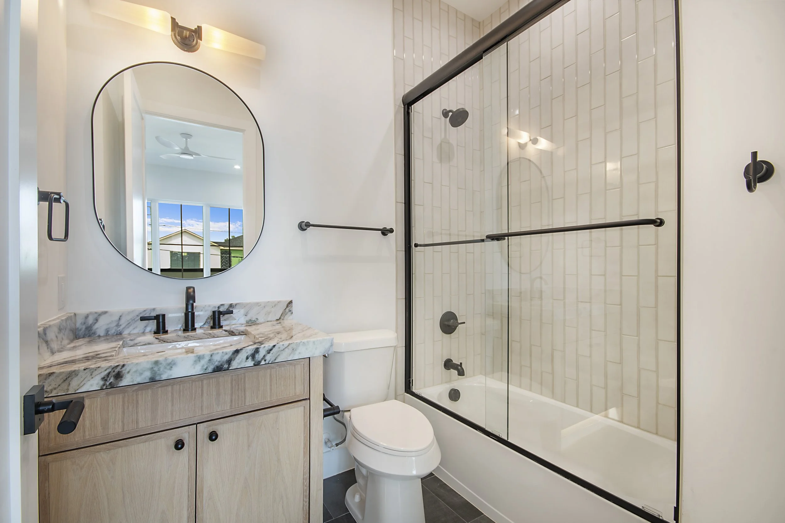 A modern bathroom with a vanity featuring a marble countertop, a round mirror, and black fixtures. There is a toilet next to a glass-enclosed shower with white tile walls and black accents. A window shows a view of a house and sky outside.