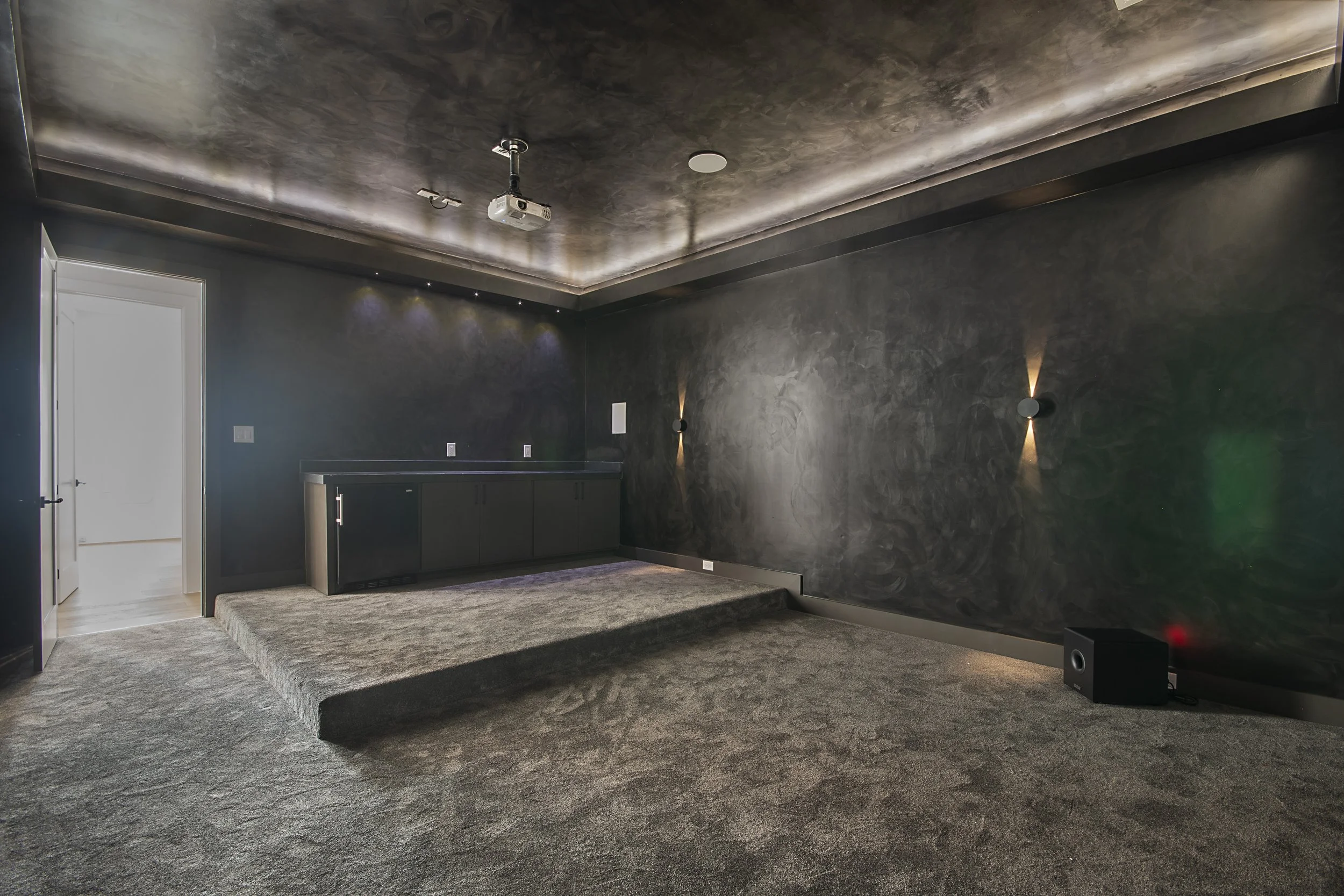 A modern home theater room with dark gray walls and ceiling, black cabinetry, a projector mounted on the ceiling, and a large speaker on the floor. The room has a carpeted floor and a small platform with a step at the front.