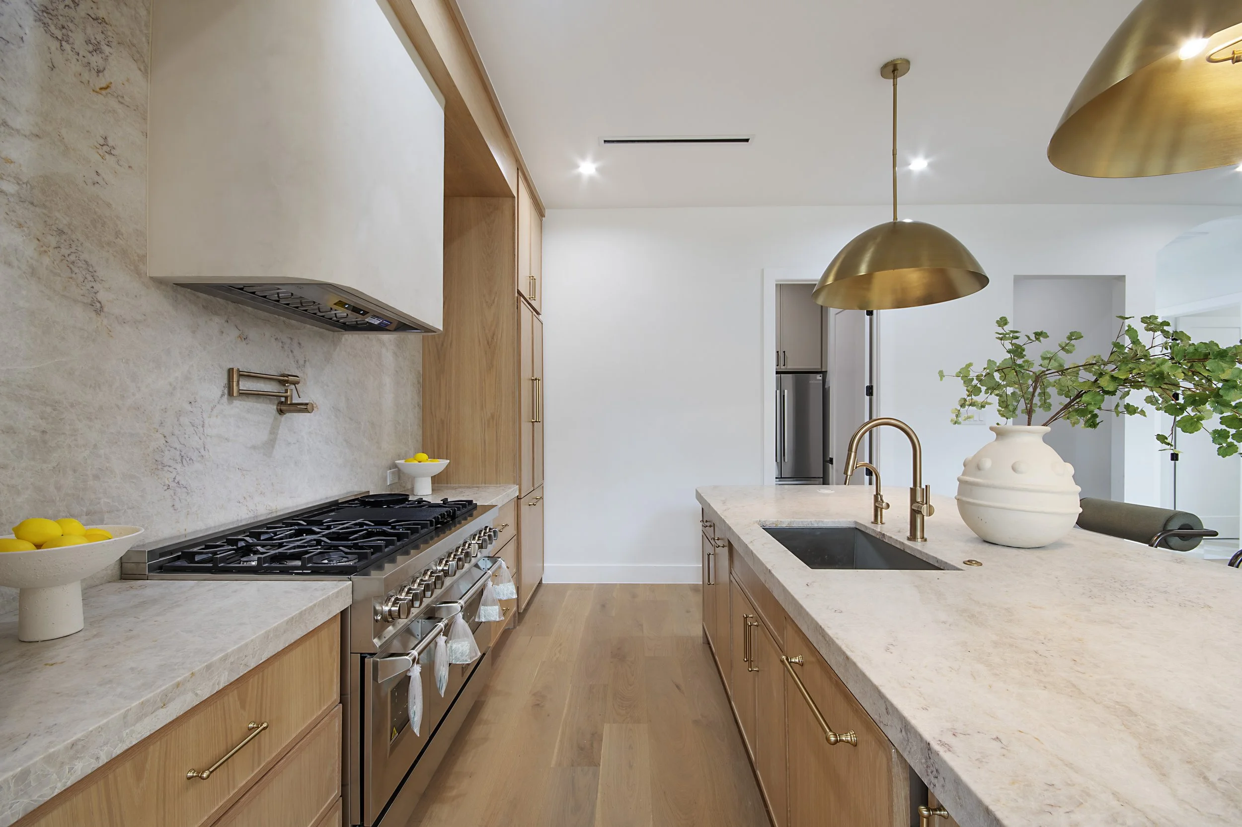 Modern kitchen with light wooden cabinets, beige marble countertops, a gas stove with oven, kitchen sink, gold fixtures, a large white vase with green foliage, and gold pendant lights.