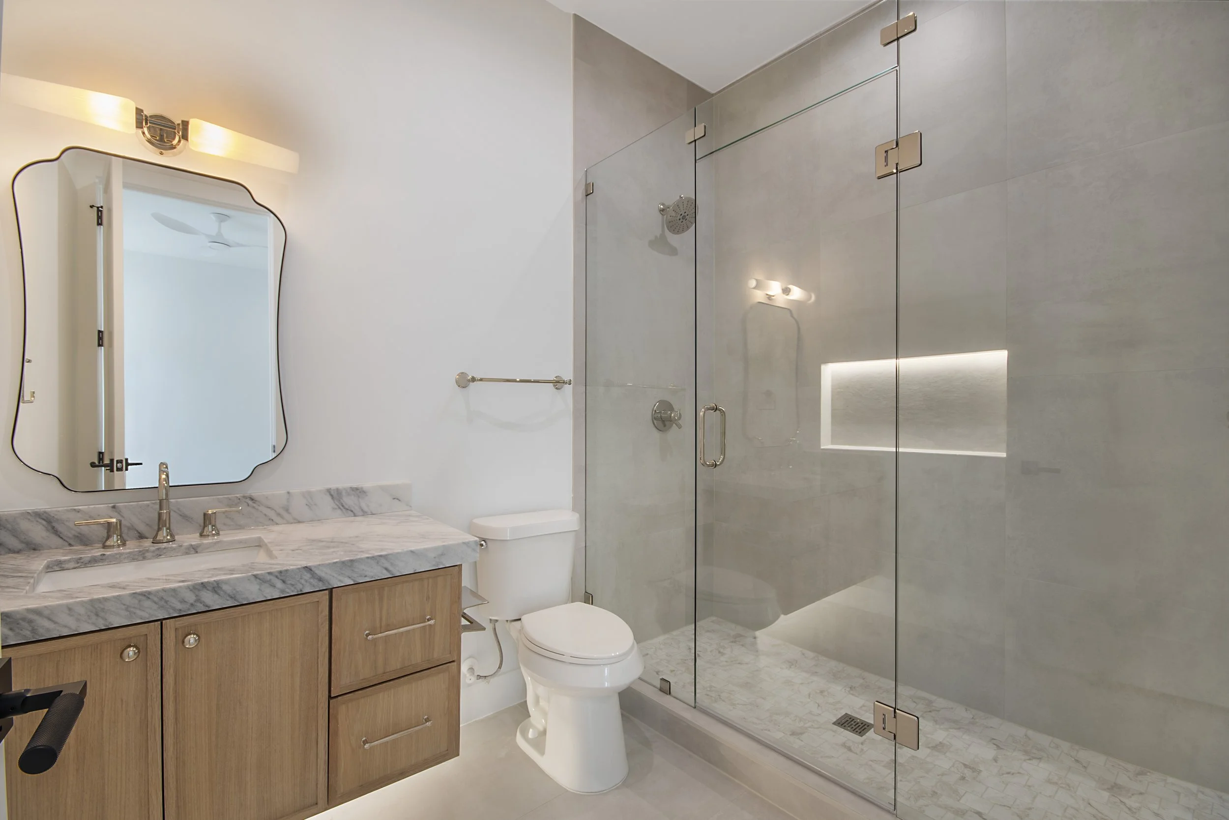 Modern bathroom with a wooden vanity, marble countertop, mirror, white toilet, and a glass-enclosed shower with built-in niche and rain shower head.