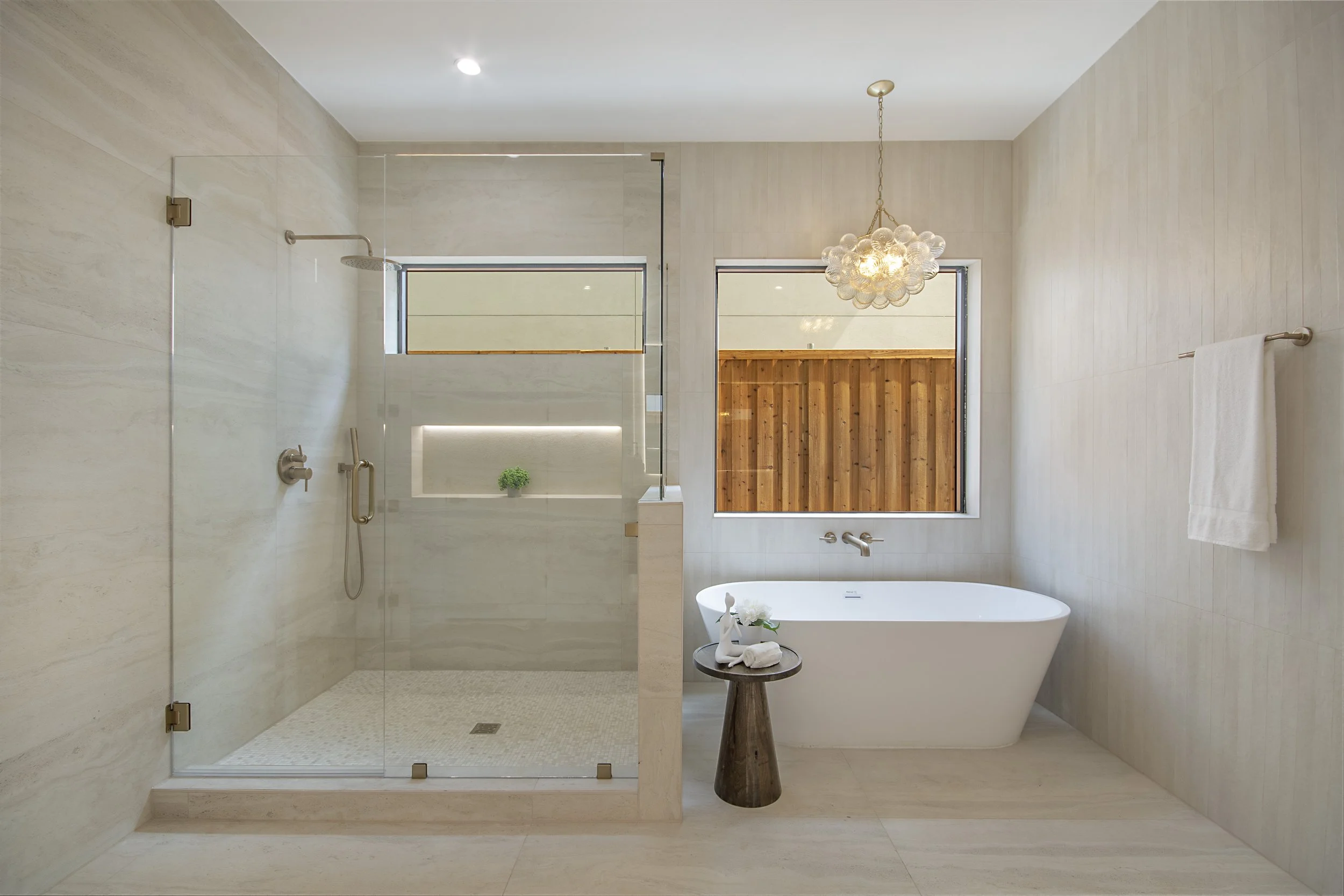 A modern bathroom featuring a glass-enclosed shower area with beige tiles, a white freestanding bathtub, a small wooden table with towels and decorative items, a chandelier hanging from the ceiling, and two windows letting in natural light.