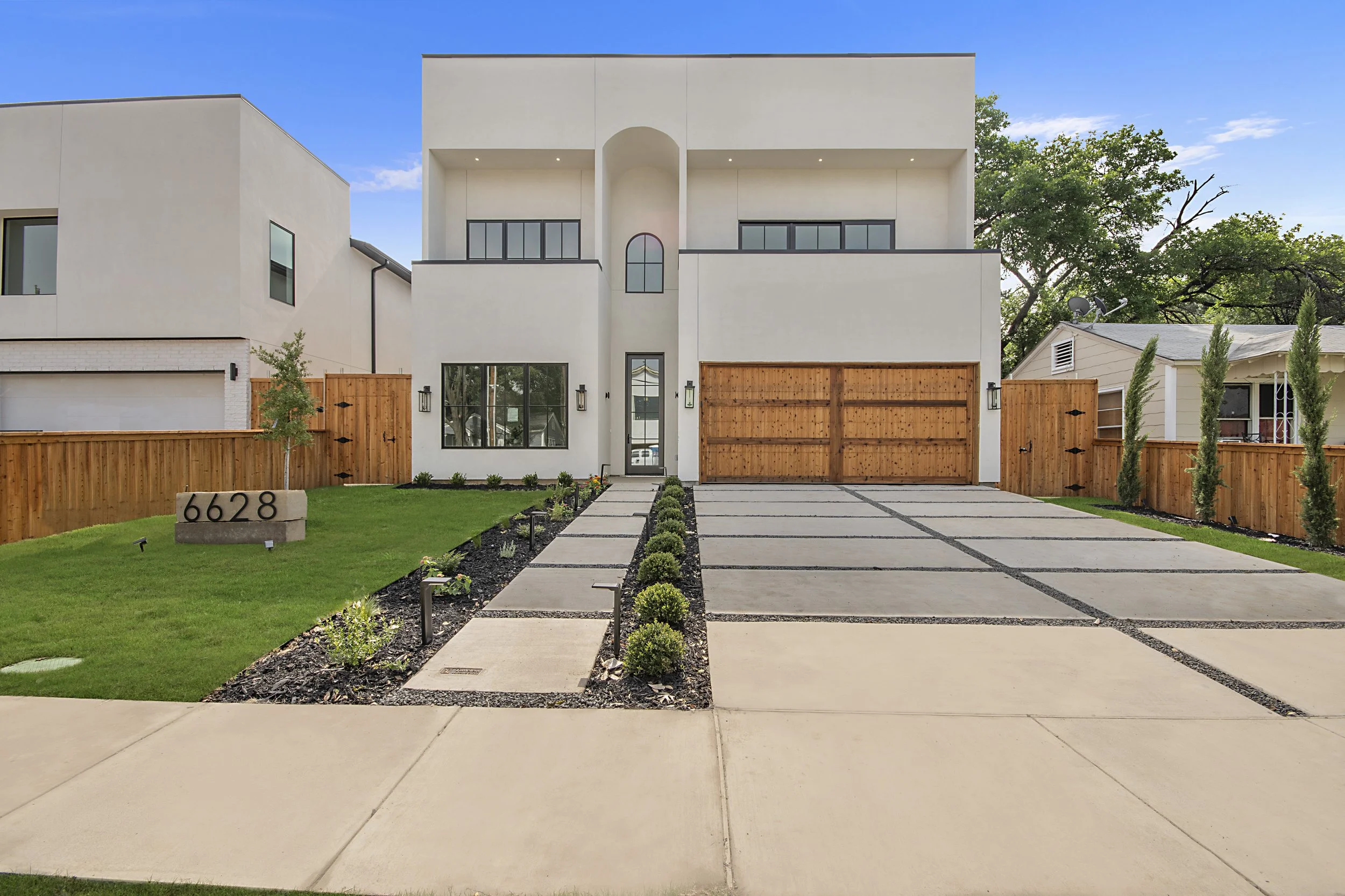 A modern white residential house with a wooden gate and fence, a concrete driveway, and a well-maintained front yard with small plants and a lawn. The house has multiple levels, large windows, and a minimalistic design. The house number "6628" is dis