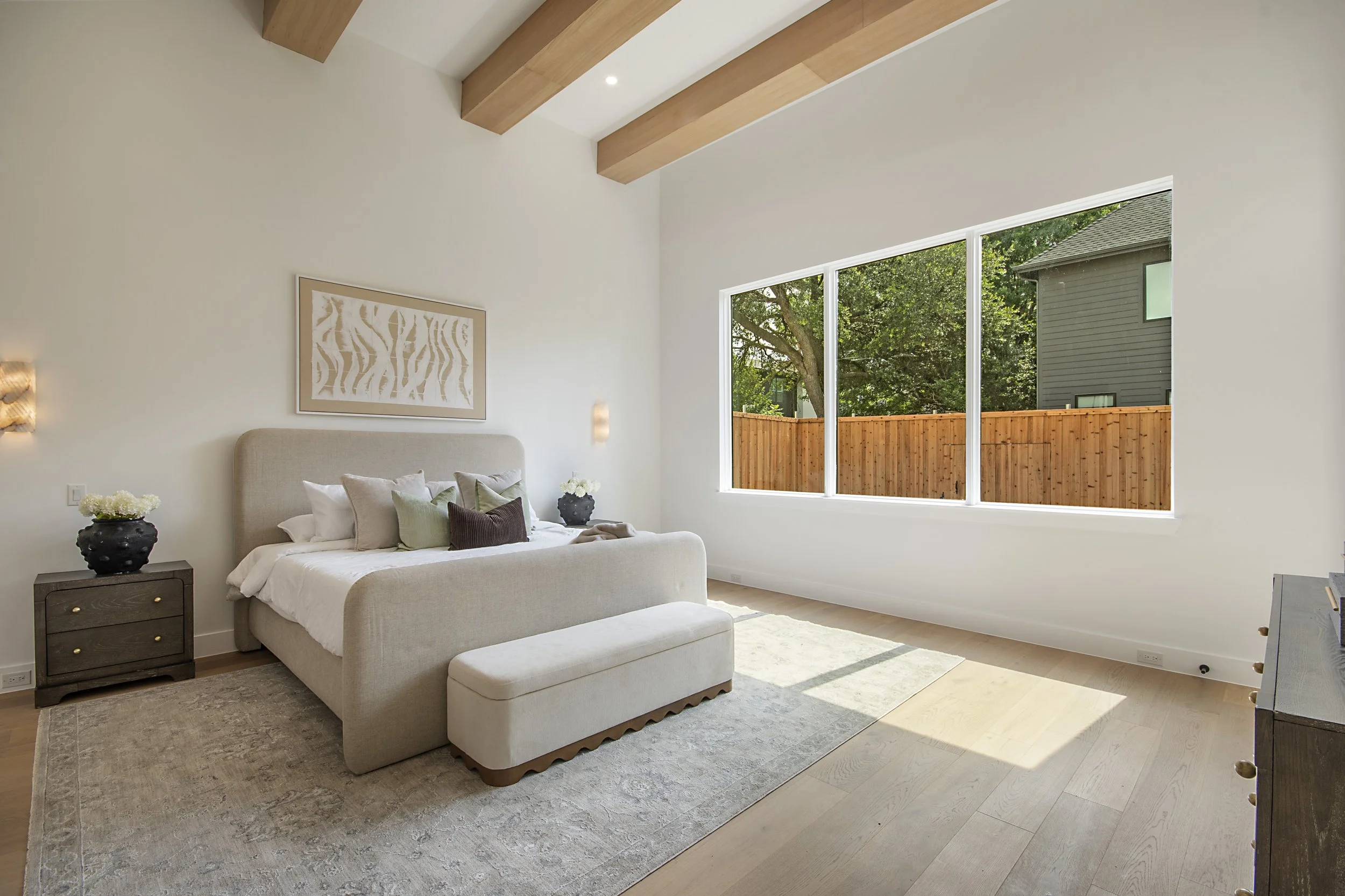 A modern bedroom with a large window, light-colored walls, a beige upholstered bed with pillows, a dark wooden nightstand with decorative vases, and a textured area rug, with wooden beams on the ceiling and trees visible outside.