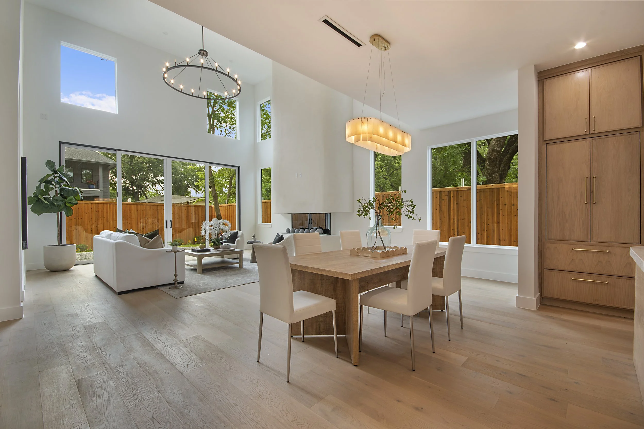 Bright open living and dining area with large windows, modern light fixtures, white walls, wood flooring, and outdoor greenery visible through the windows and sliding glass door.