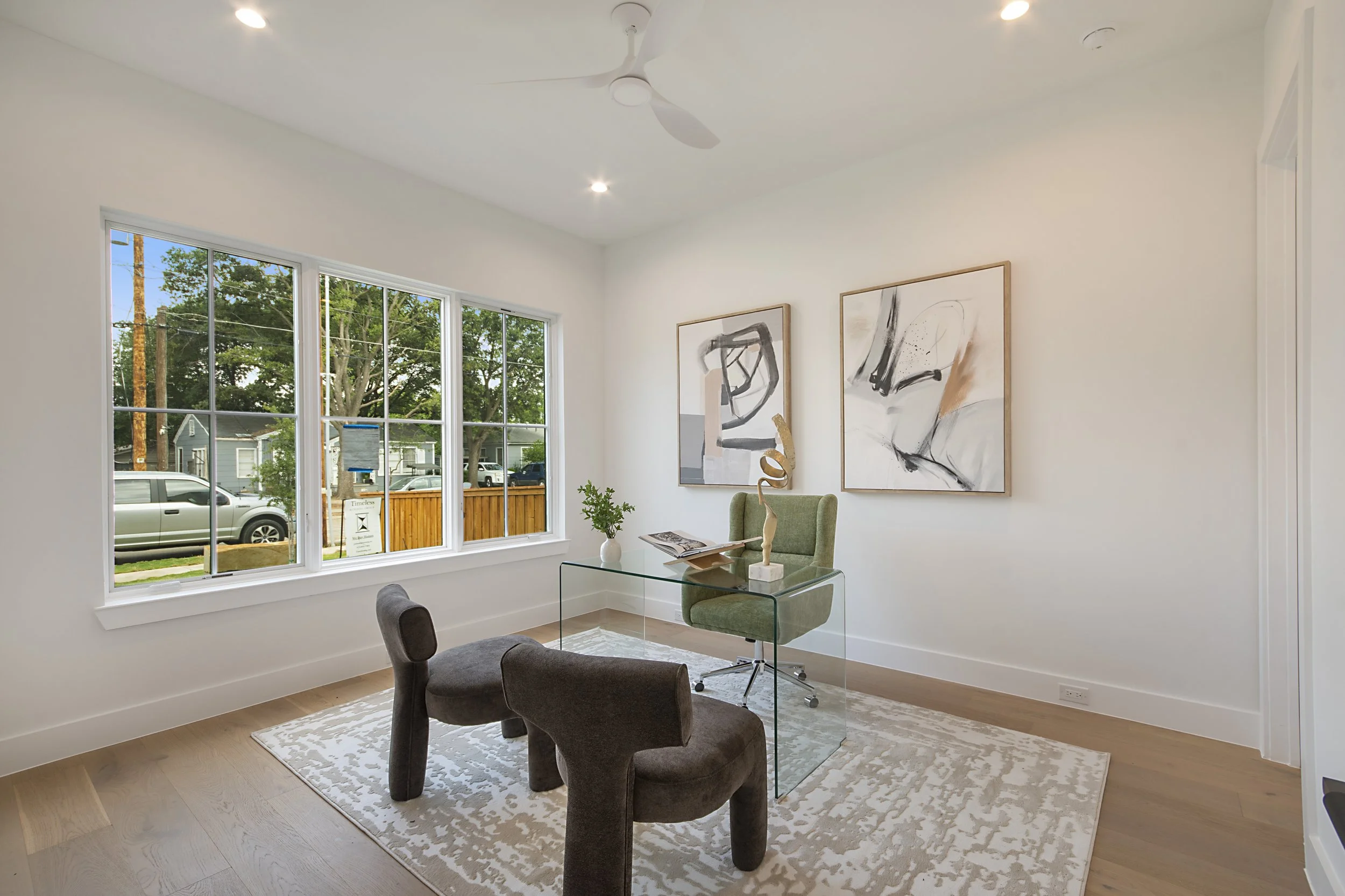 A modern living or office room with a large window, abstract black and white artwork on the wall, a green chair behind a glass desk with decorative objects, and a patterned rug on wooden flooring.