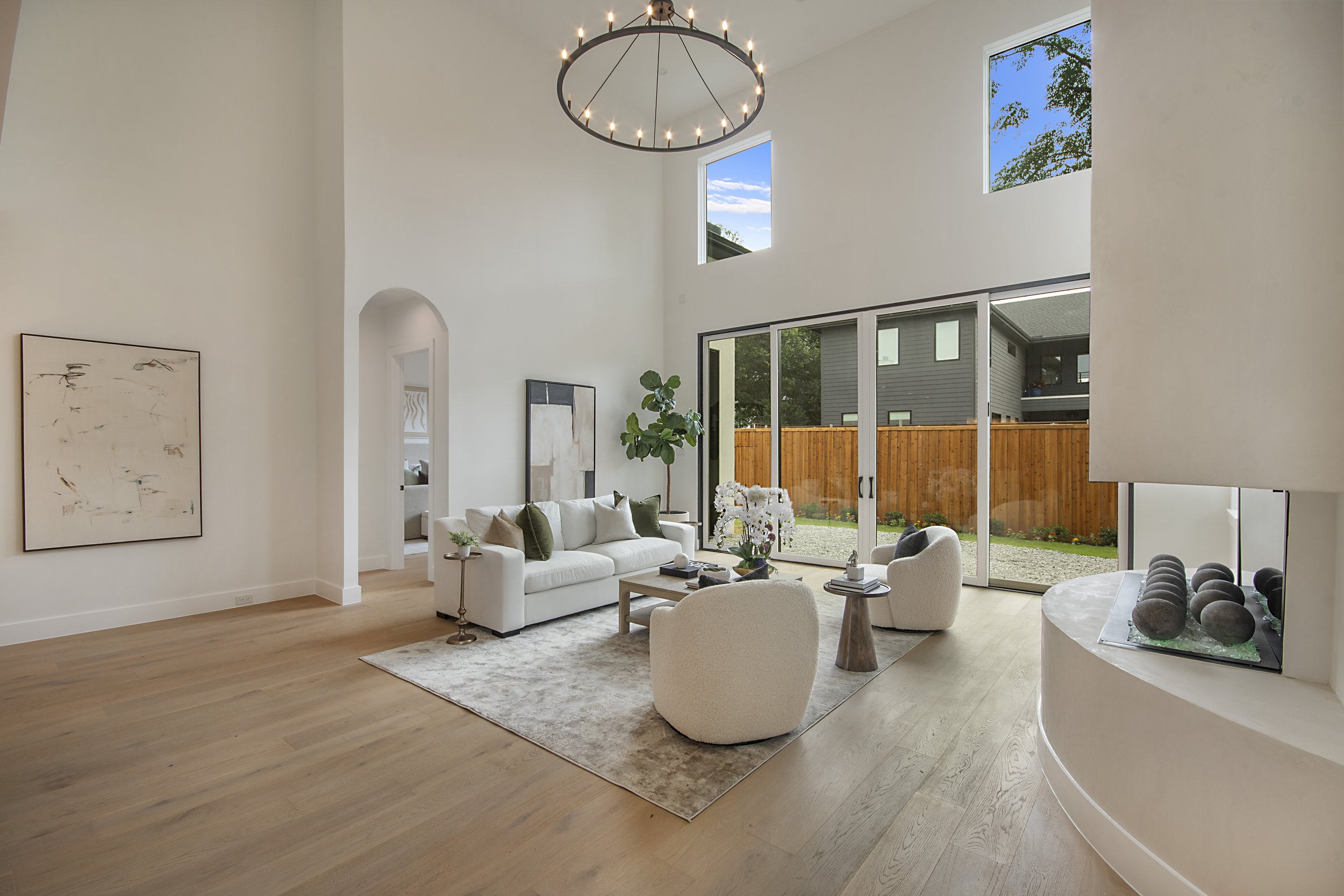 Spacious living room with high vaulted ceiling, large windows, white walls, and light wooden flooring. Features a white sofa with pillows, a large abstract painting on the wall, a round coffee table with decorative items, and a potted plant near slid