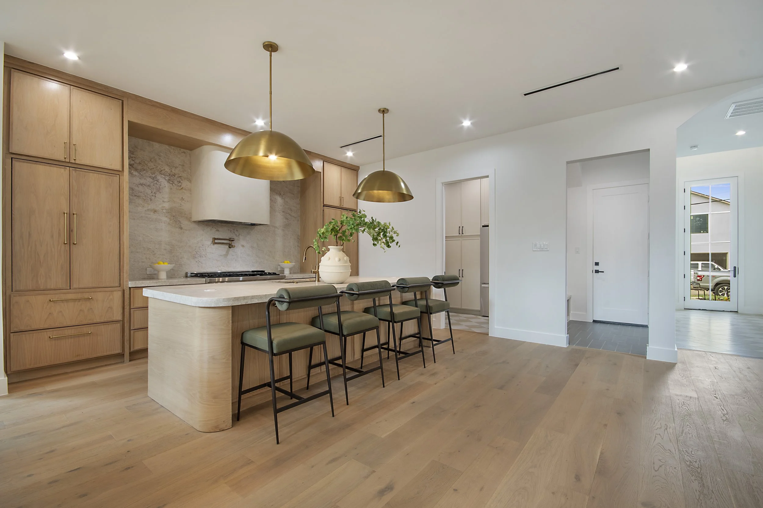 Modern open-concept kitchen with light wood cabinetry, a white kitchen island, green bar stools, gold pendant lights, a white vase with greenery on the island, and a door with a window in the background.