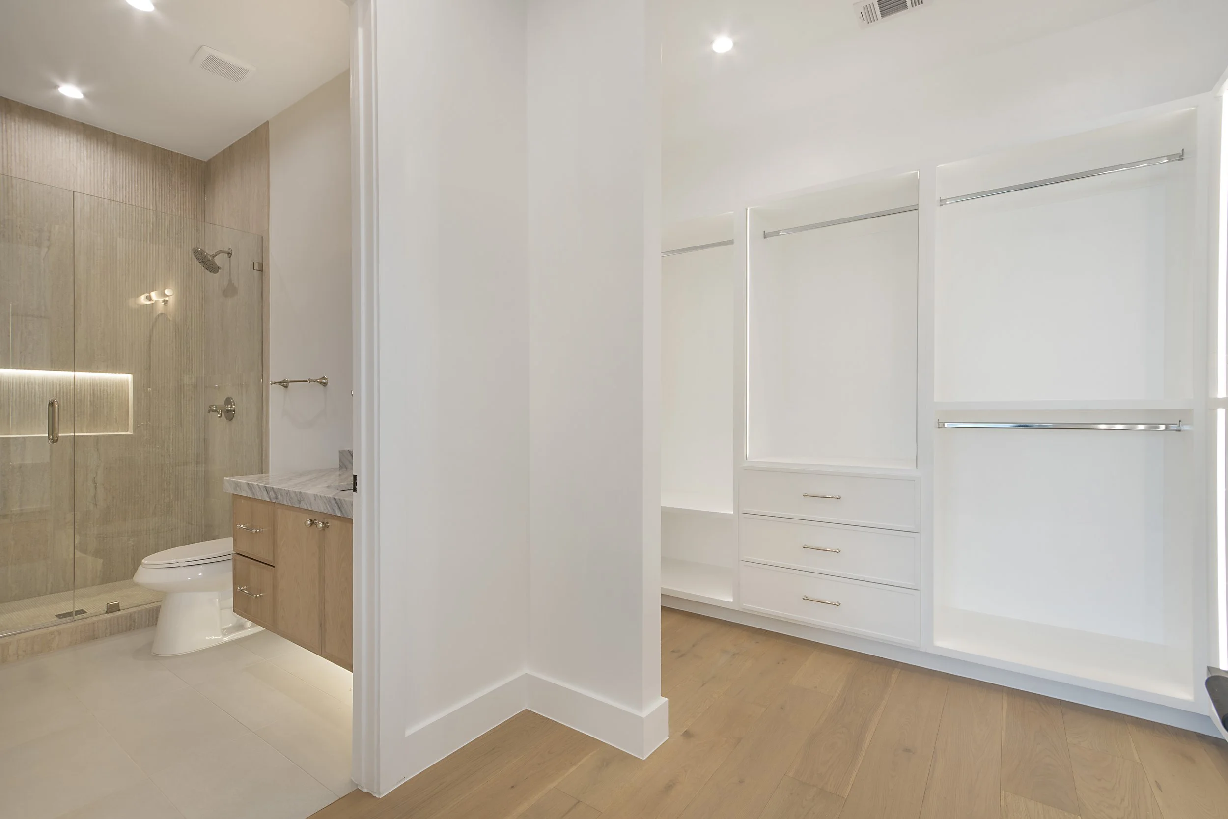 Modern bathroom with a glass-enclosed shower, a white toilet, a wooden vanity with a marble countertop, and white built-in cabinets on wood flooring.