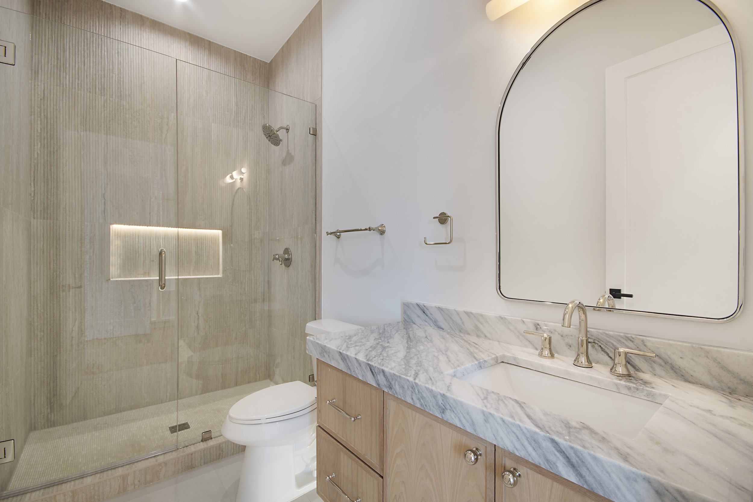 Modern bathroom with a glass-enclosed shower, a white toilet, a marble countertop with a sink, a large mirror, and light-colored wooden cabinets.