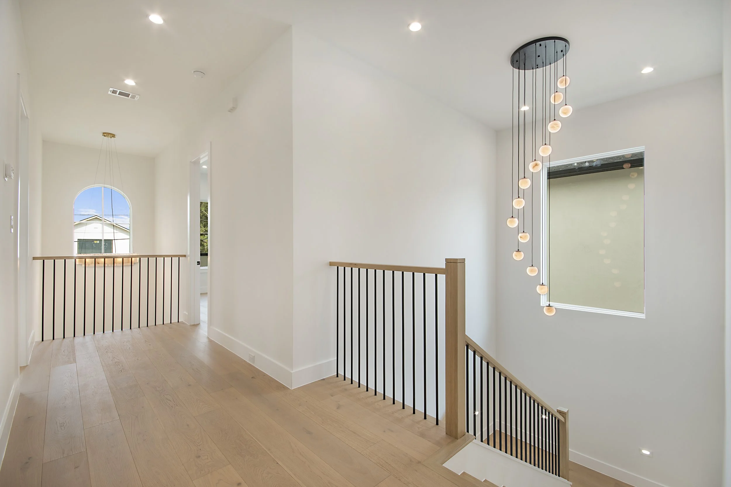 Interior view of a modern, bright staircase landing with white walls, light wood flooring, black and wood railing, and a high ceiling. Features a distinctive hanging chandelier with multiple spherical lights.
