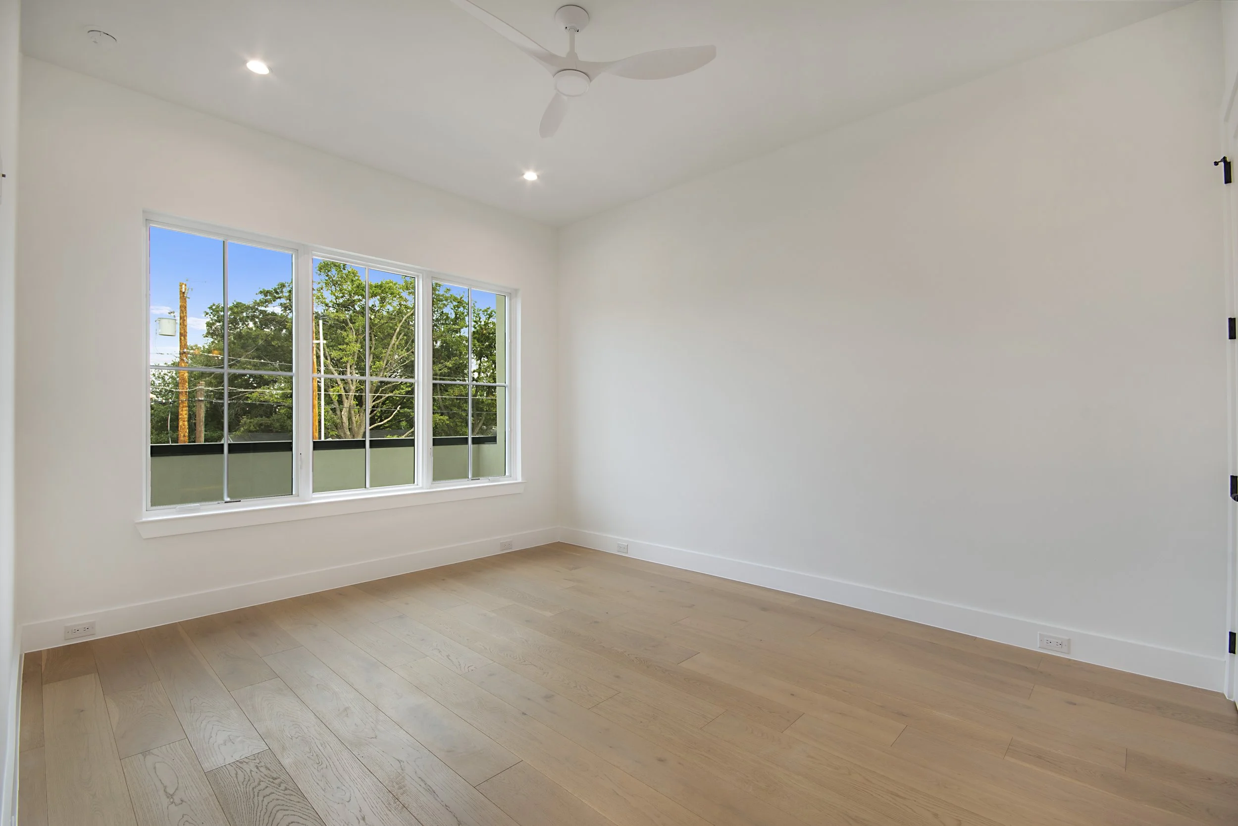 An empty room with white walls, a large window showing trees outside, recessed ceiling lights, a white ceiling fan, wood flooring, and electrical outlets.