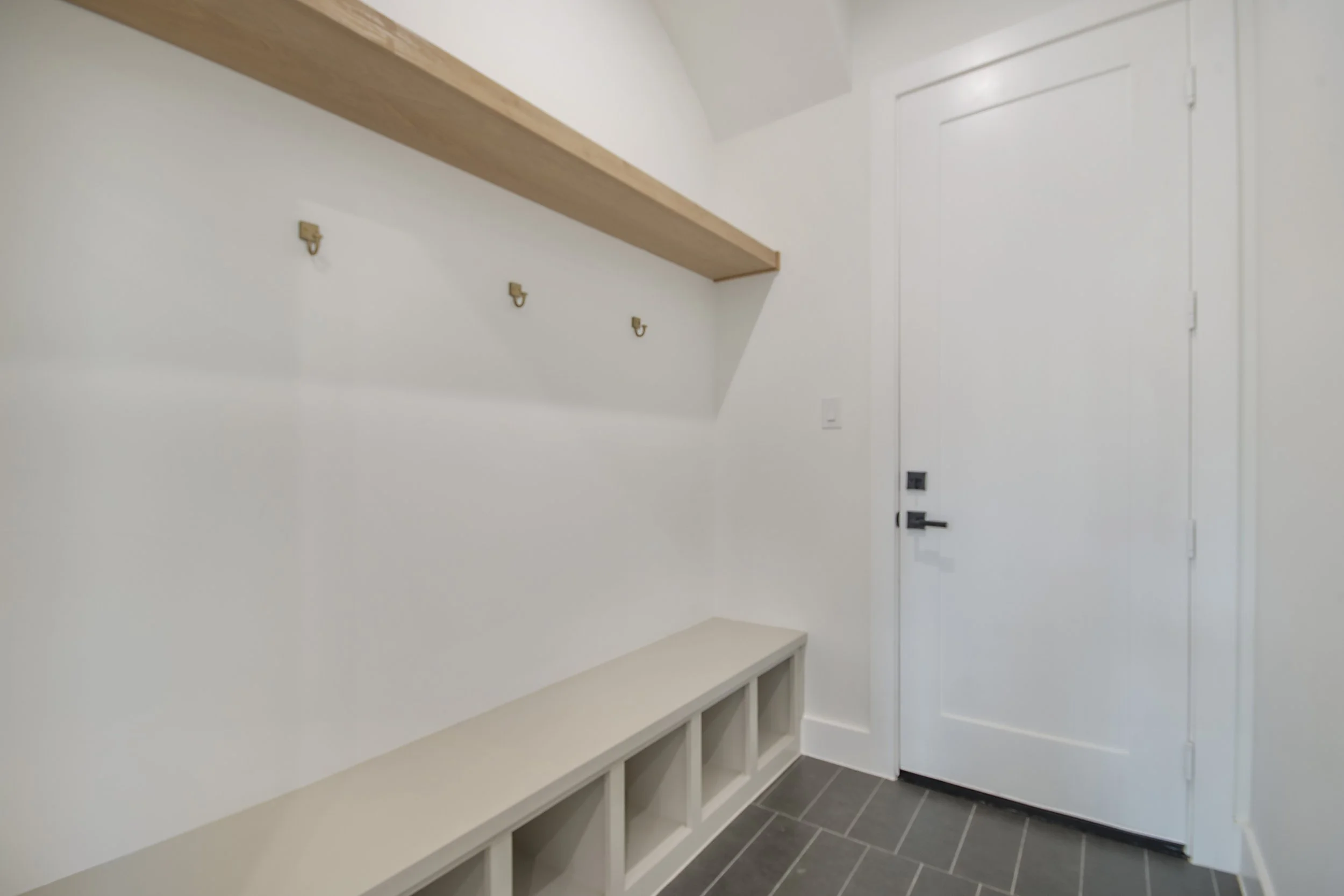 Entryway with white walls, using hooks, a wooden shelf, and a bench, near a white door with a black handle.