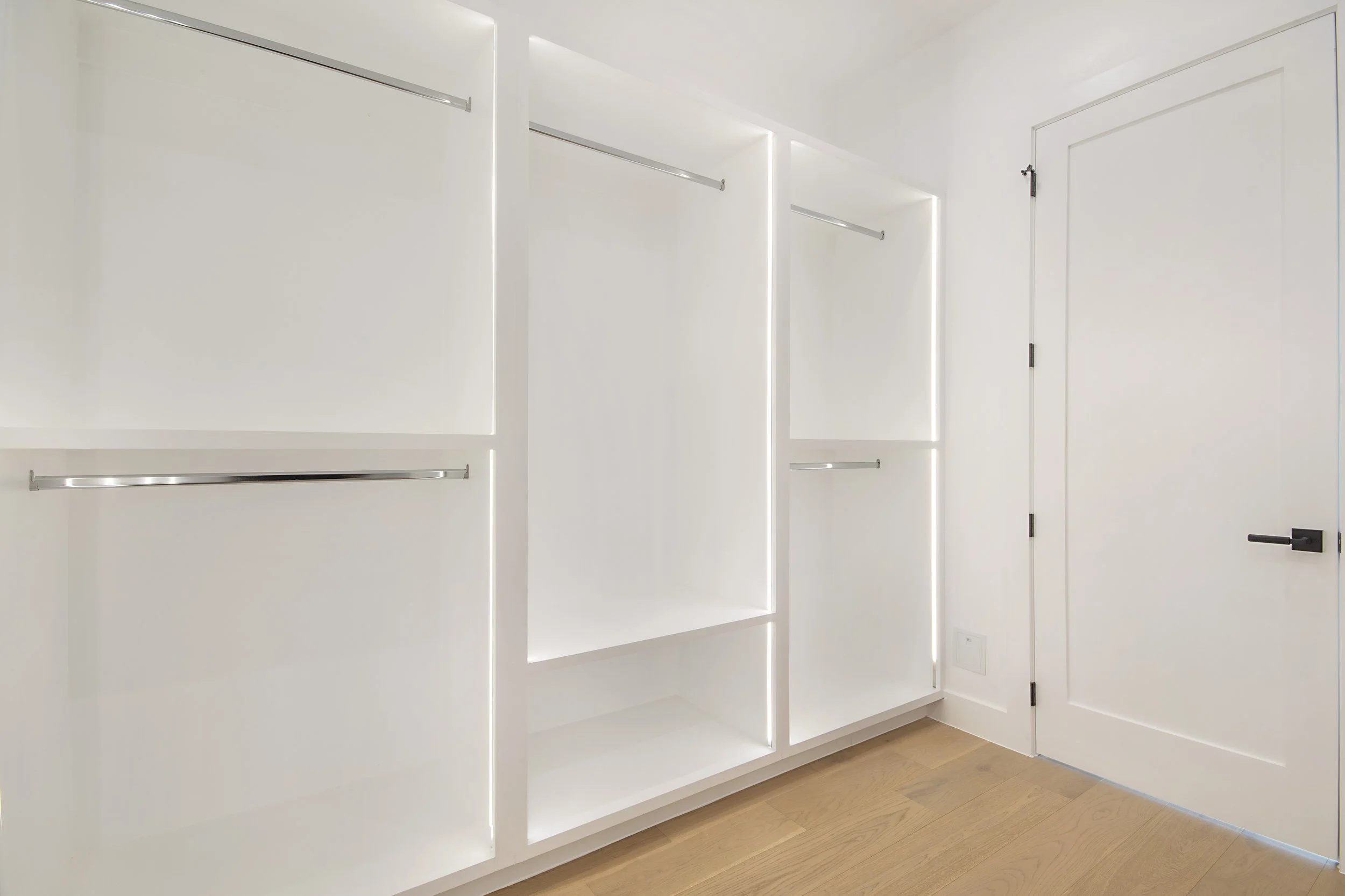 Empty white storage closet with open shelves and hanging rods, next to a closed white door in a room with light wood flooring.