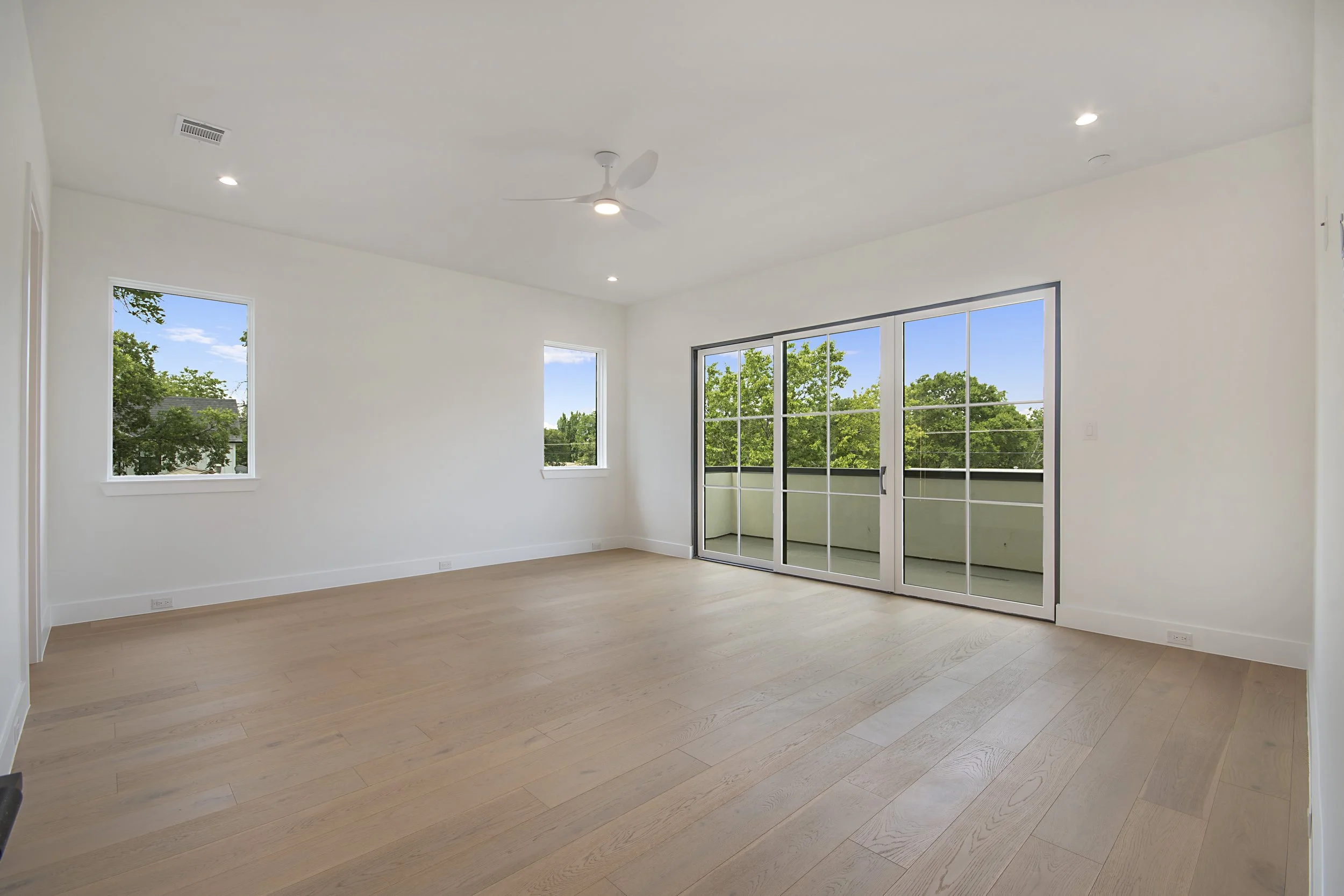 Empty living room with light wood flooring, white walls, large windows showing trees, and a sliding glass door leading to a balcony.