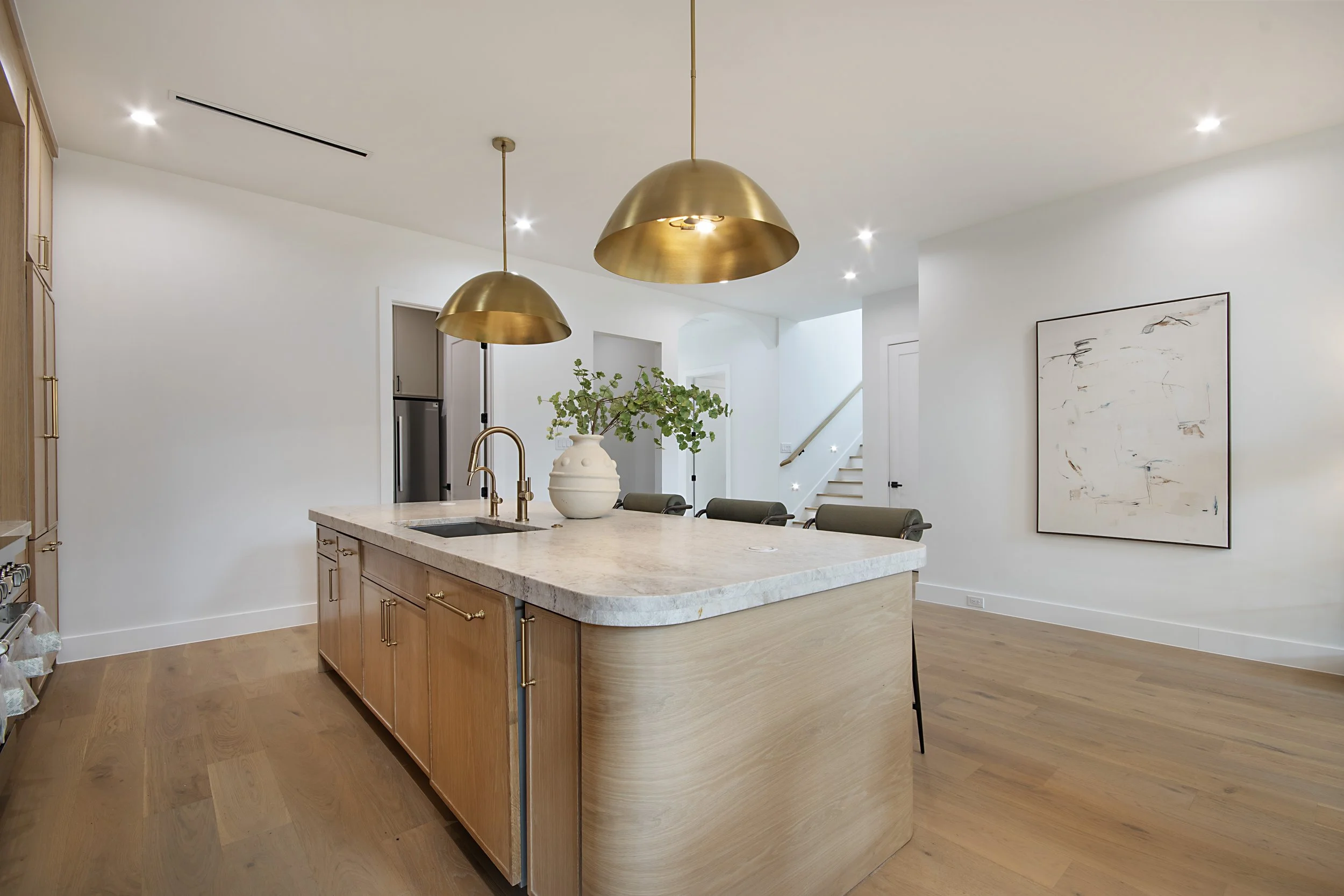 Modern kitchen interior with a large kitchen island, gold pendant lights, white walls, wooden cabinets, and a wooden floor. There are chairs at the island and artwork on the wall.