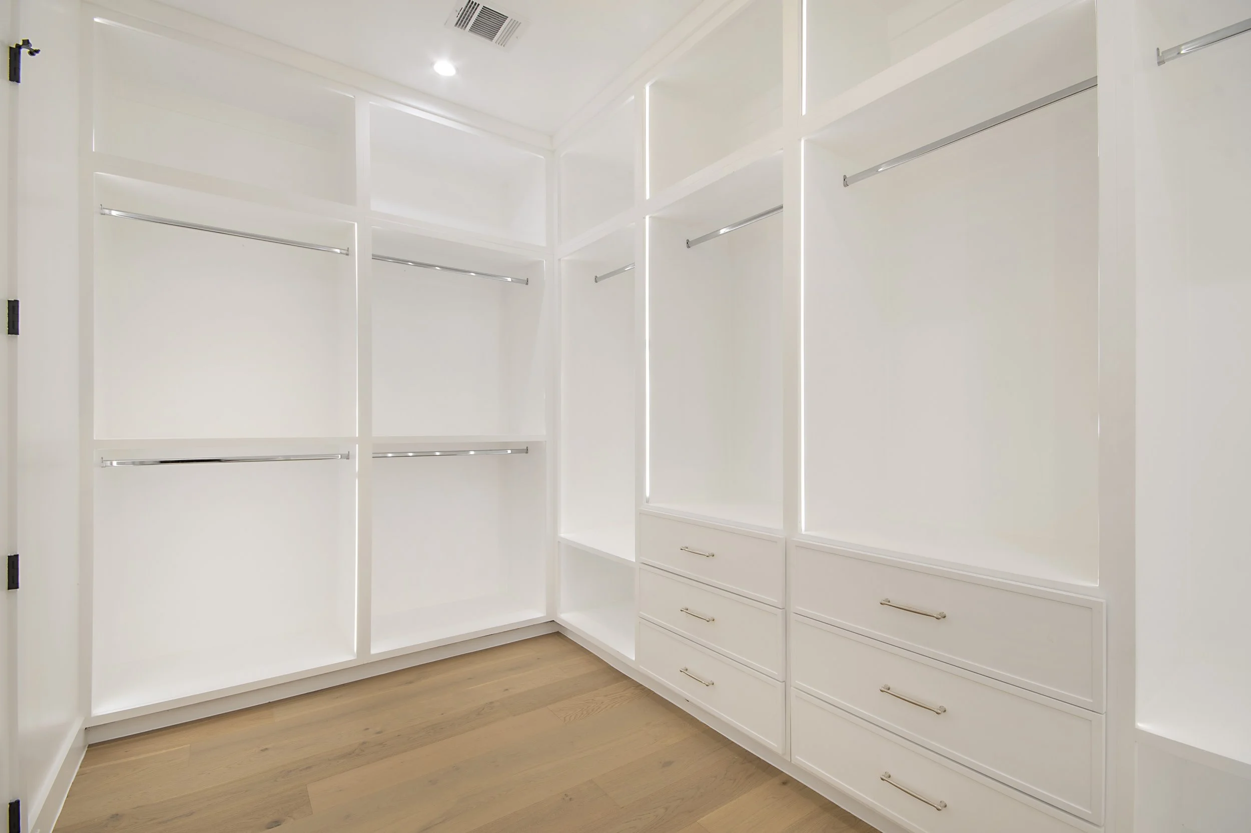 Empty white walk-in closet with built-in shelves, drawers, and hanging rods, hardwood floor, and ceiling light.