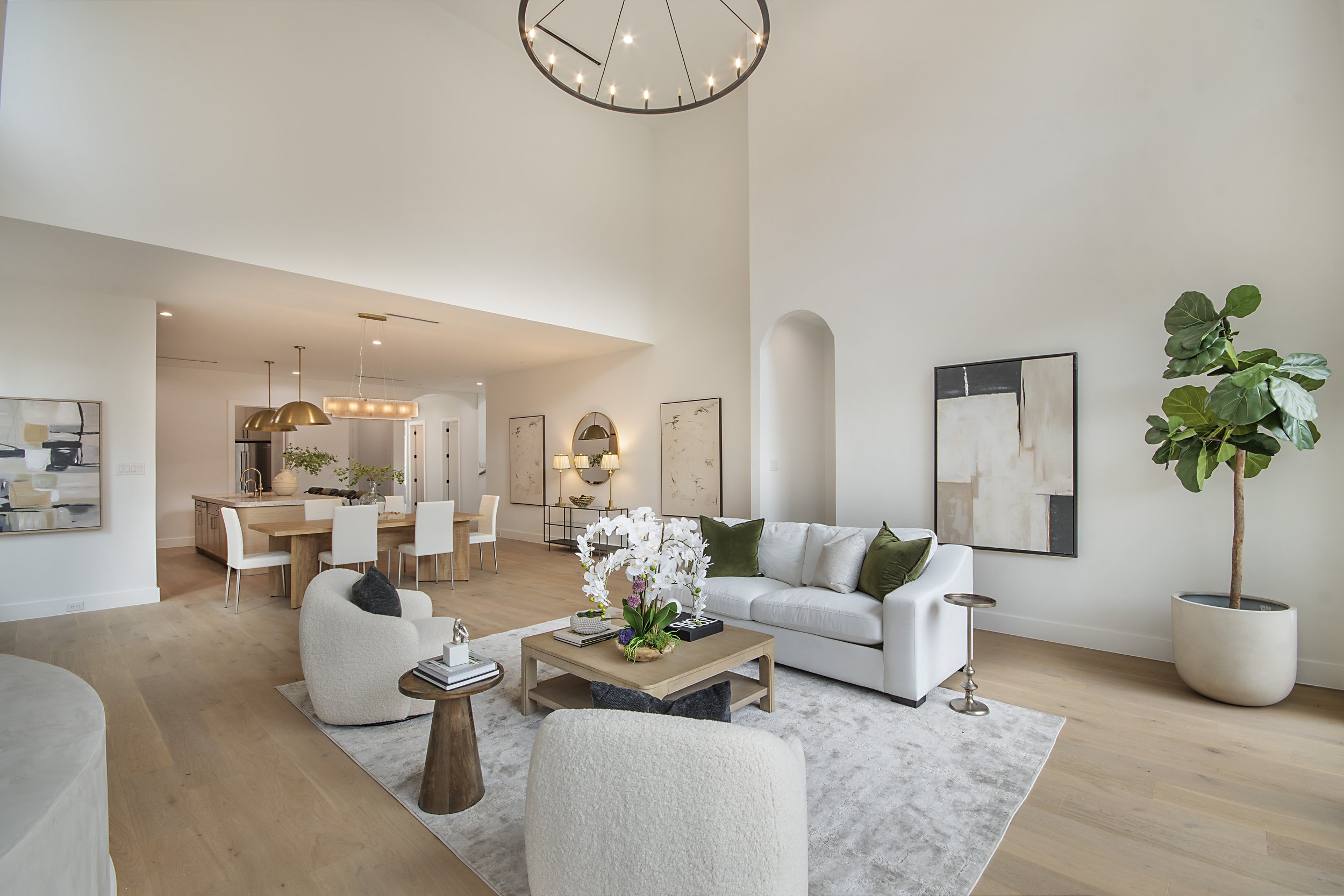 Living room with white walls, modern artwork, a white sofa with green and beige cushions, a coffee table with orchids, an armchair, and tall indoor plants, illuminated by contemporary lighting fixtures.
