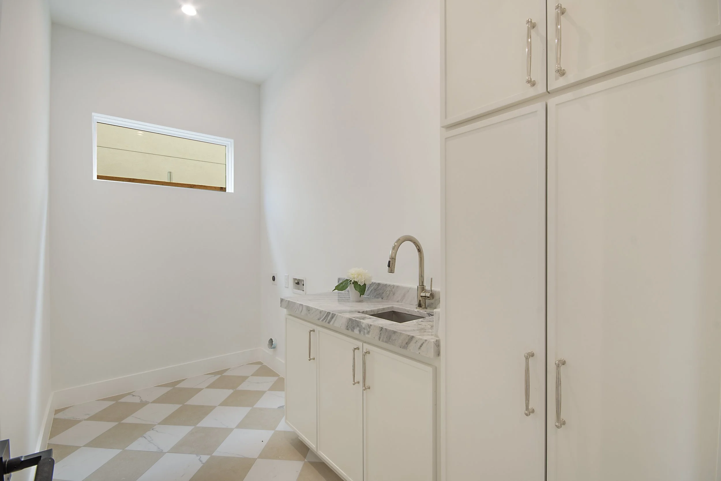 A small kitchen with white cabinets, marble countertop, and a stainless steel faucet. The wall has a small window, and the floor has a checkered pattern of beige and white tiles.