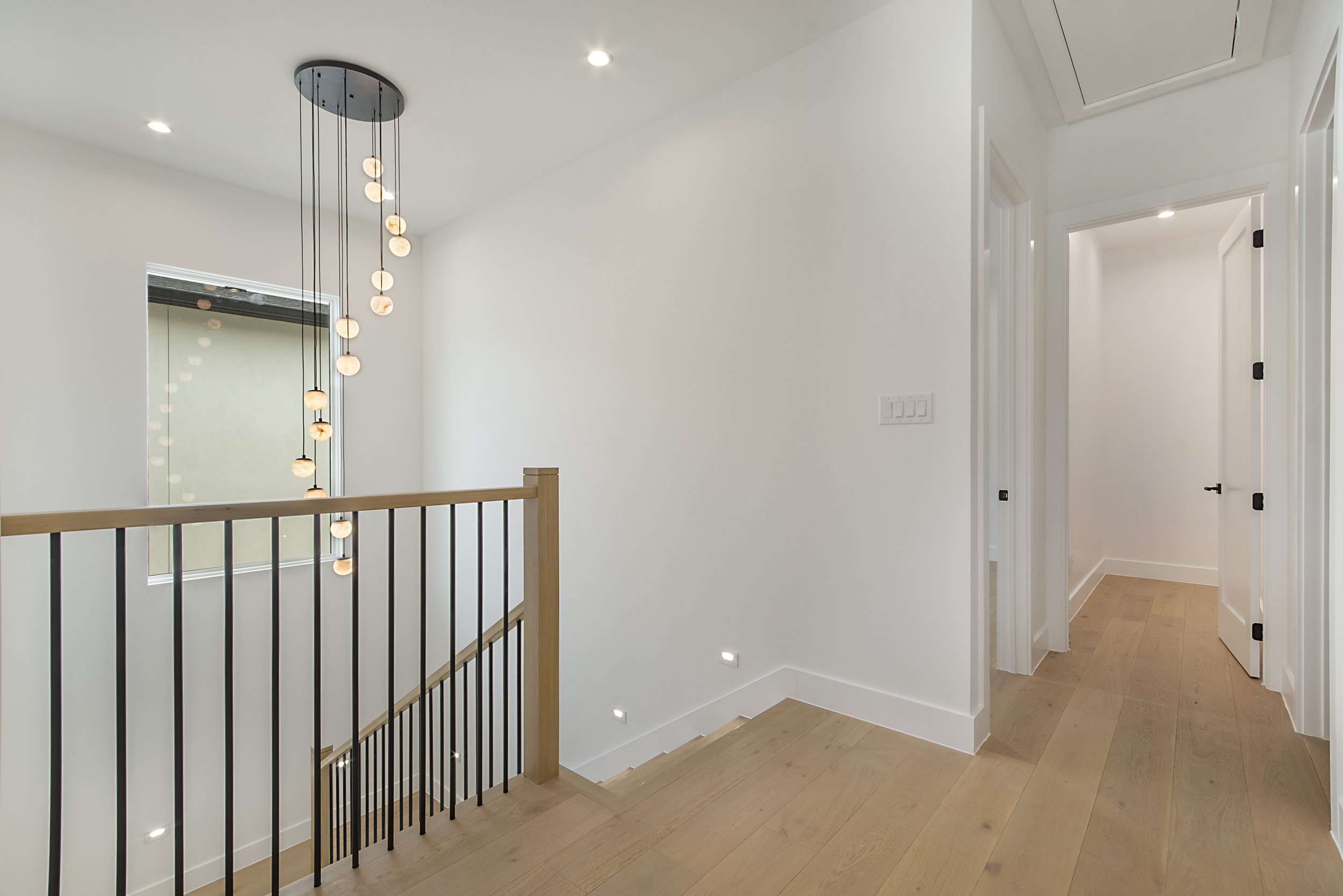 Interior view of a modern home hallway with white walls, light wooden flooring, several open doors, recessed ceiling lights, and a decorative hanging light fixture.