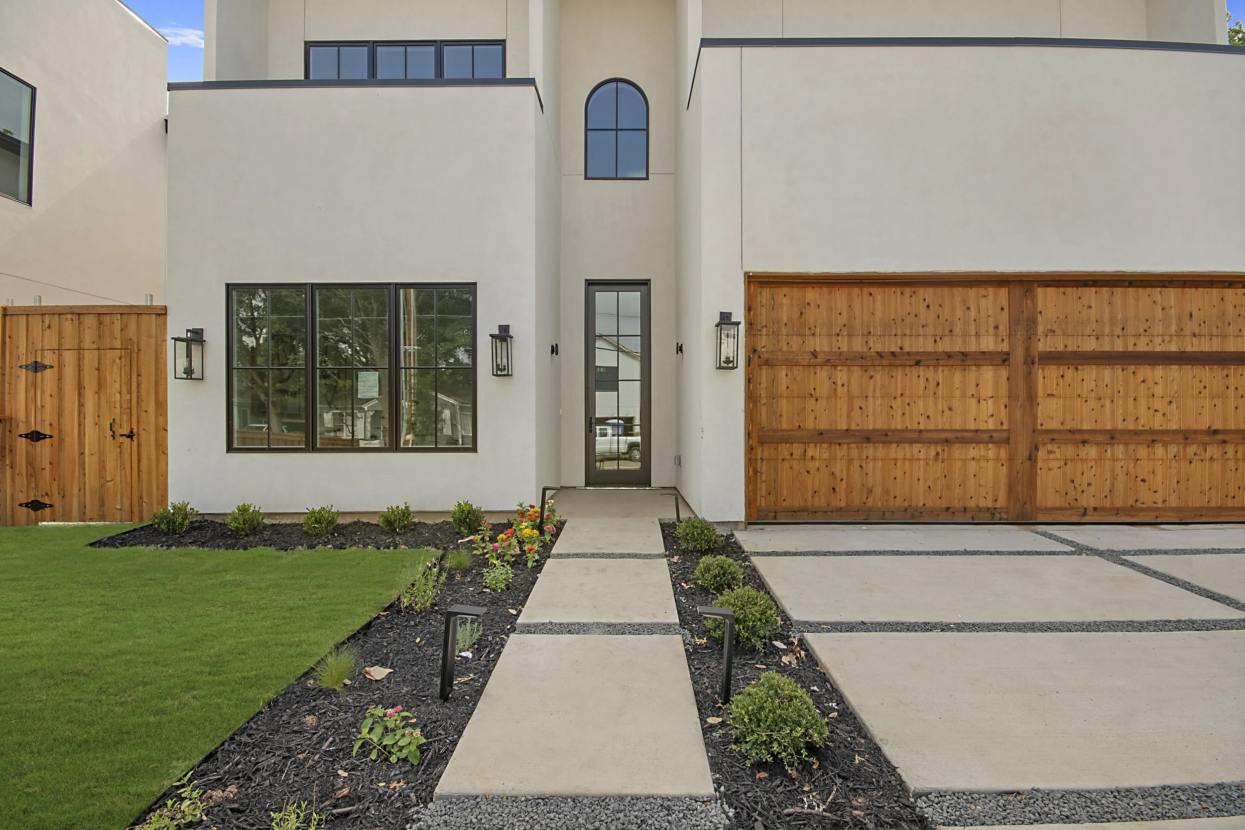 Front view of a modern residential house with a white exterior and wooden garage door. It has a concrete pathway leading to the front door, flanked by small plants and flowers, and a well-maintained lawn.