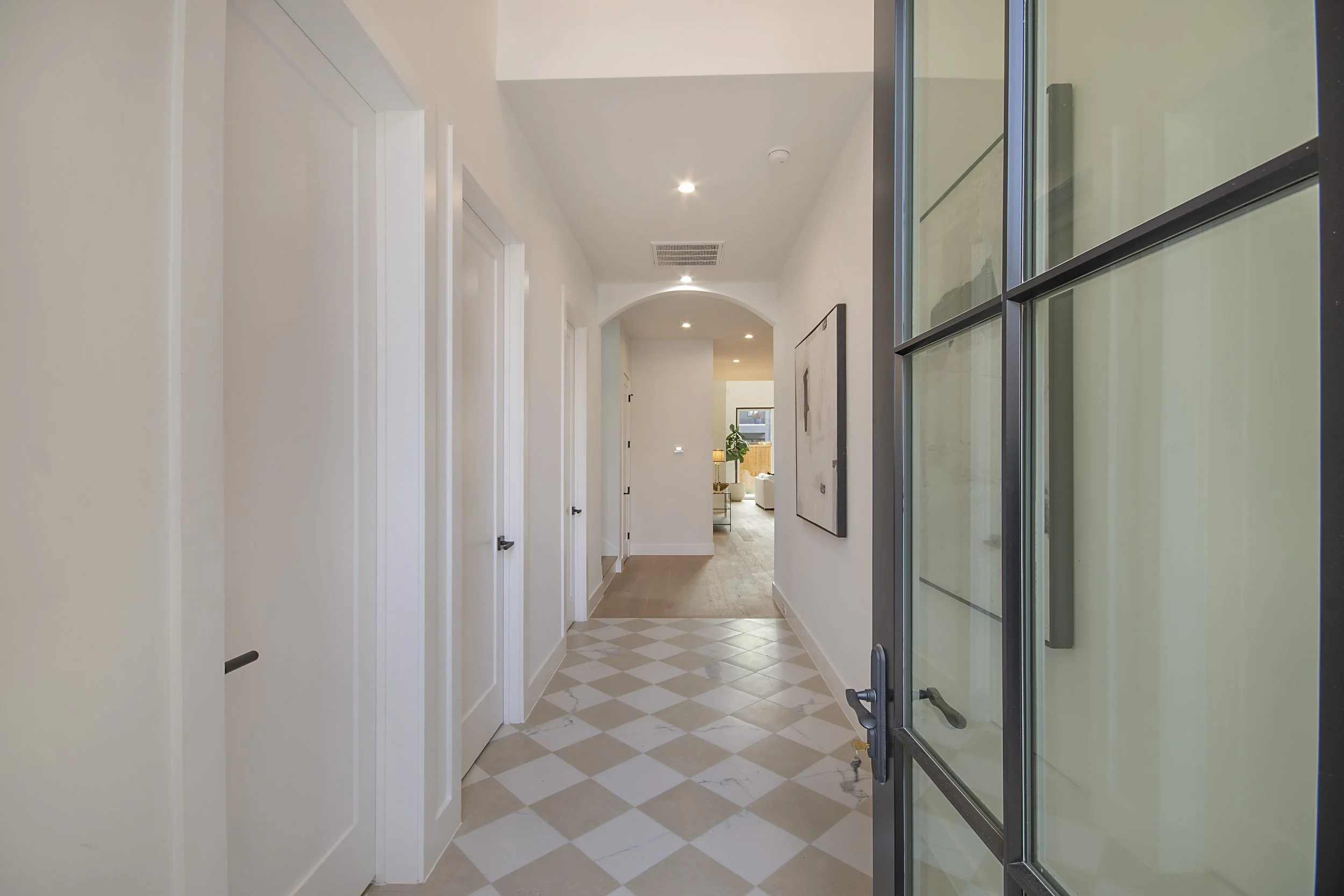 A hallway with white walls and doors, diamond-patterned tile flooring, and recessed ceiling lights. A glass door is partially visible on the right, and a framed picture hangs on the wall. In the background, a living area with a lamp and plants can be