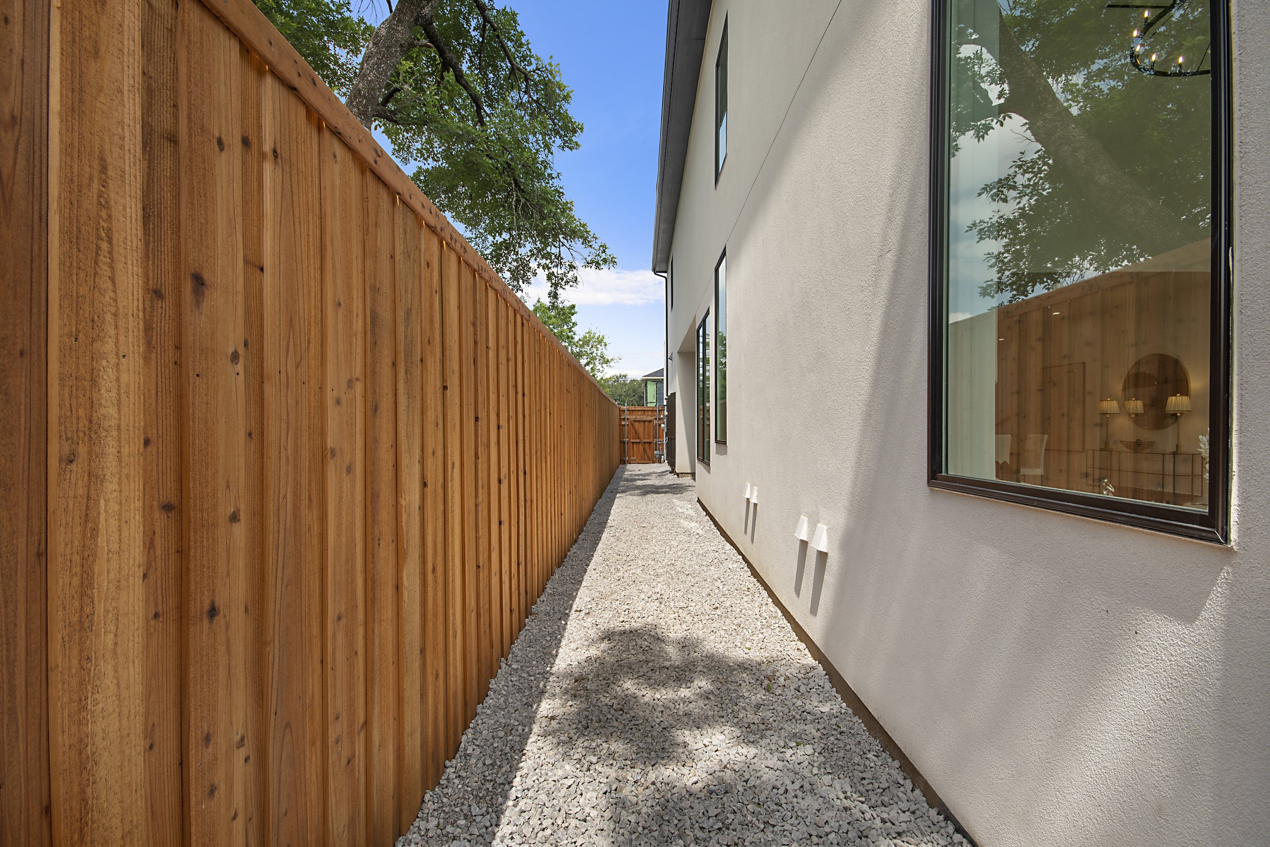 Side yard between a wooden fence and a white modern house with large windows, gravel ground, and trees overhead on a sunny day.