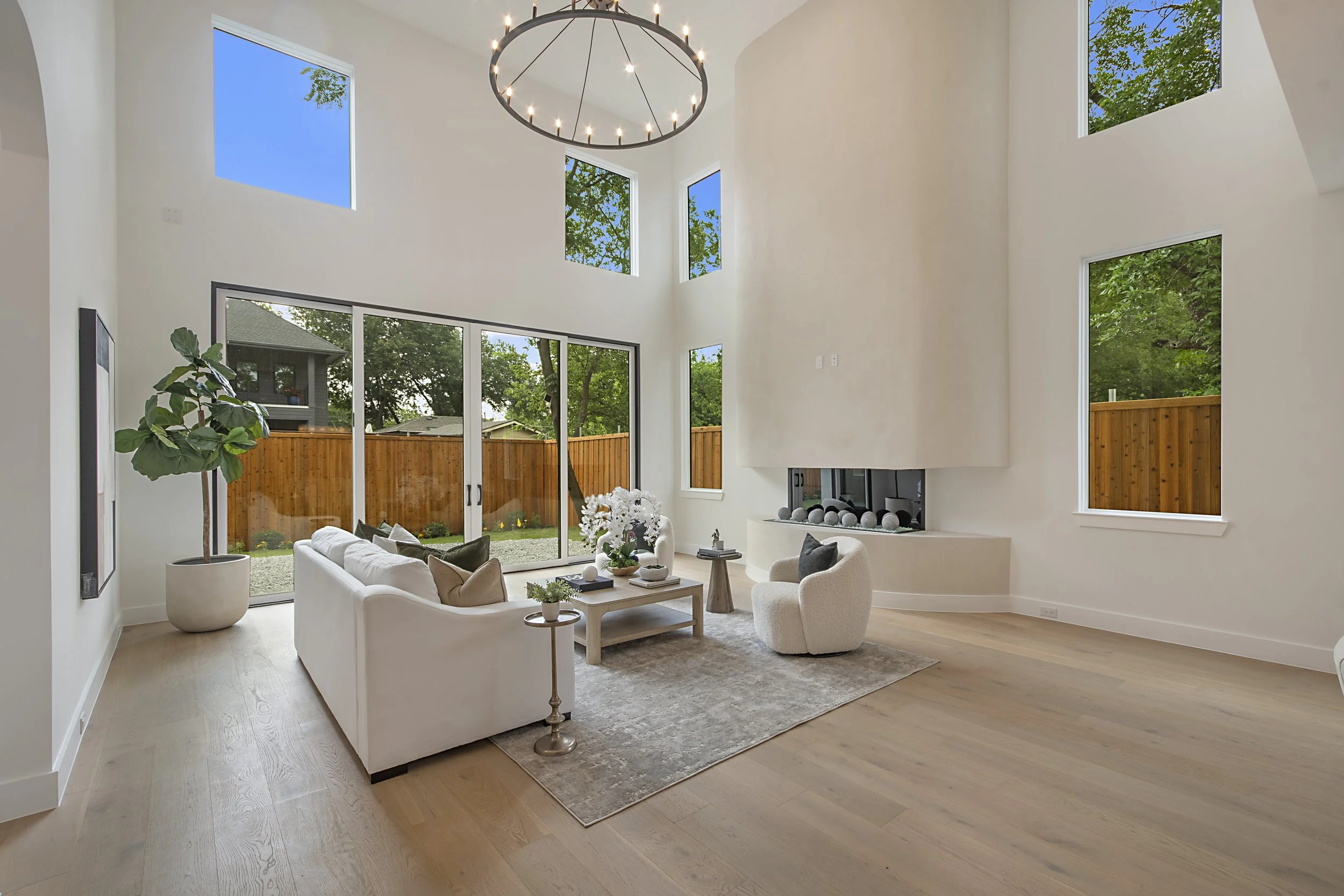 Bright living room with a high ceiling, large windows, and modern decor. Features a white sofa, armchair, coffee table, and a large potted plant. The windows reveal a blue sky, trees, and a backyard with a wooden fence. A circular chandelier hangs fr