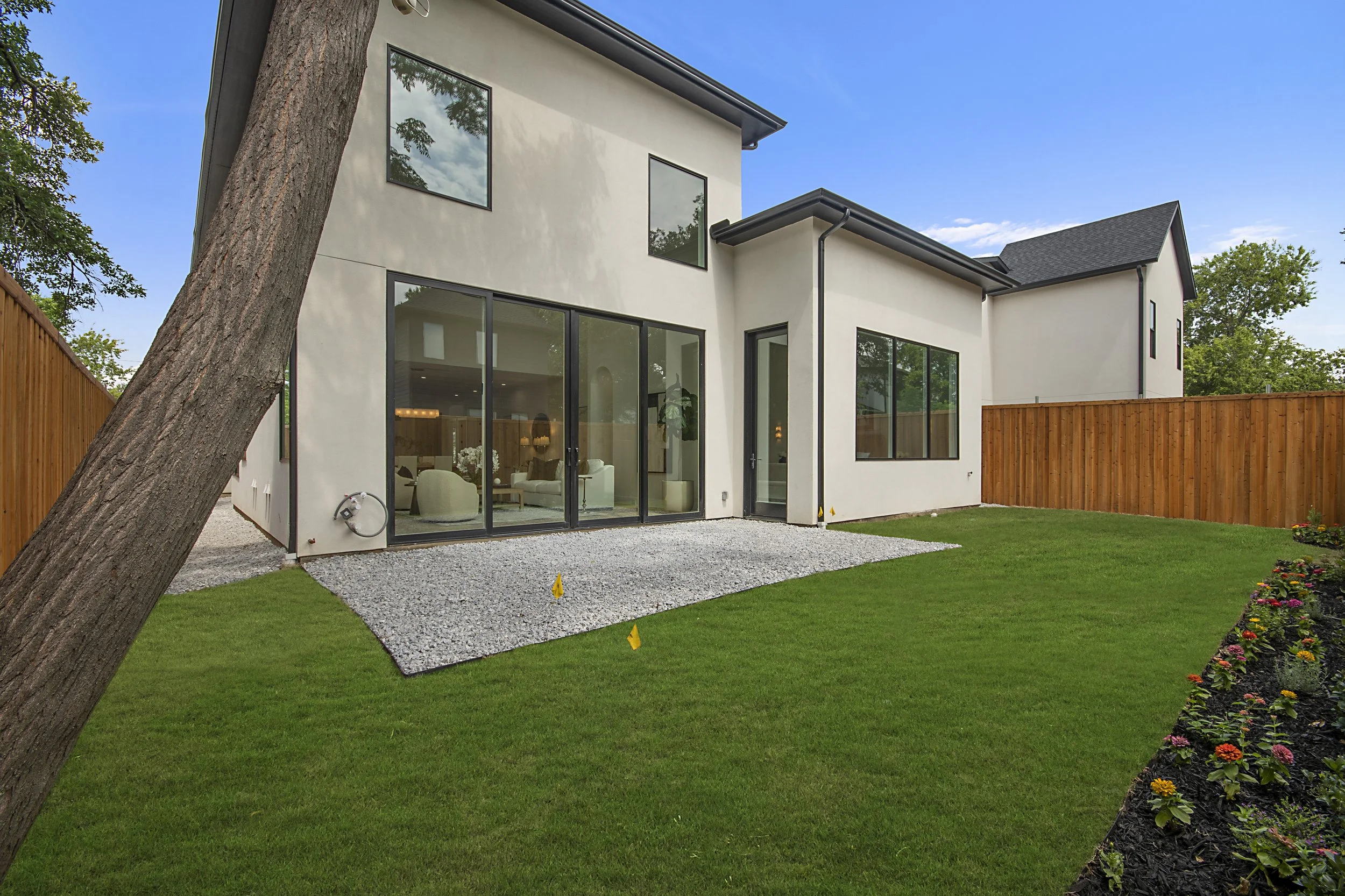 Modern two-story house with large glass doors and windows, surrounded by a wooden fence, a manicured lawn, and flower beds under a blue sky.