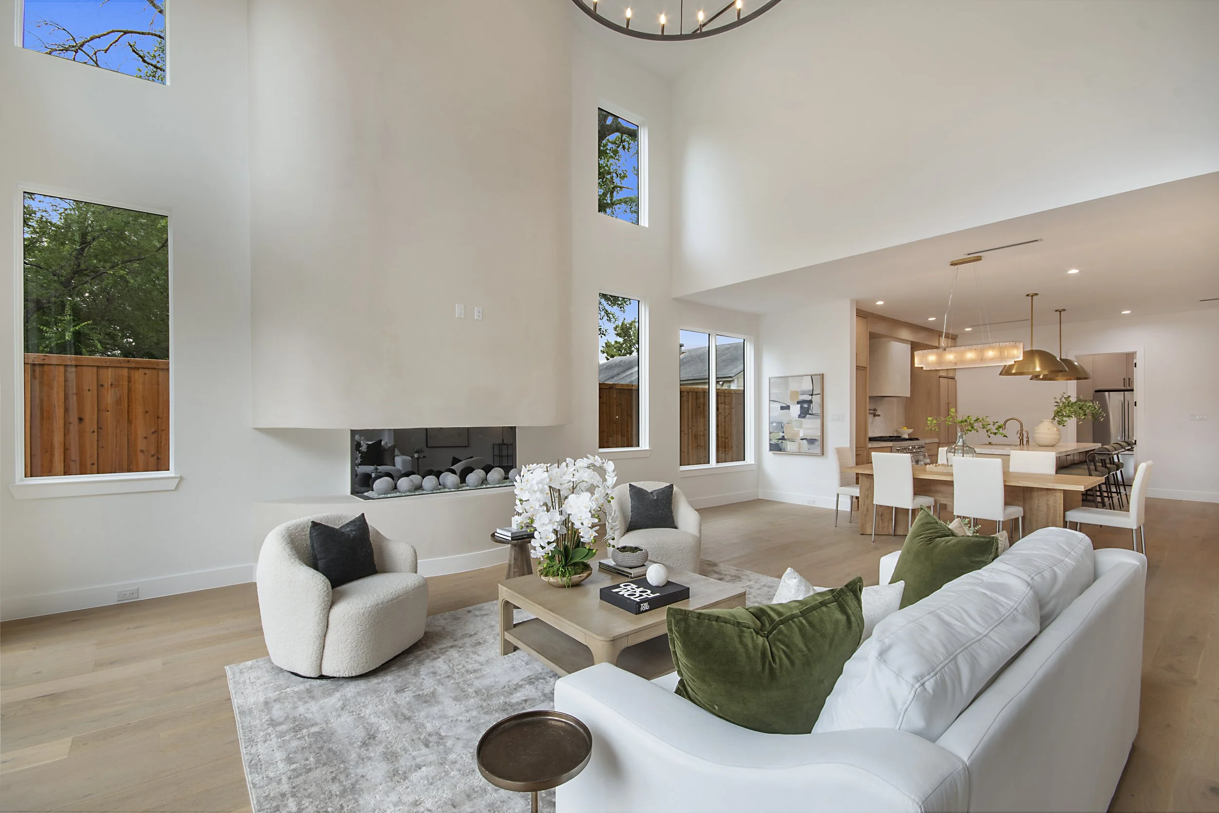 A bright, modern living room with high ceilings, large windows, and a fireplace. The room is decorated with cozy furniture including white and green seats, and a wooden coffee table with books and a plant. Sliding glass doors lead to an outdoor space