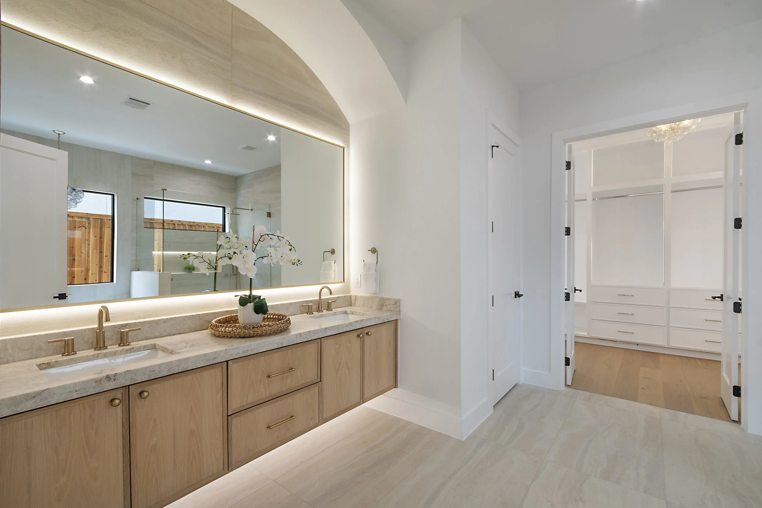 A modern bathroom with a double sink vanity, large mirror, orchid plant, and a walk-in closet with white cabinetry.