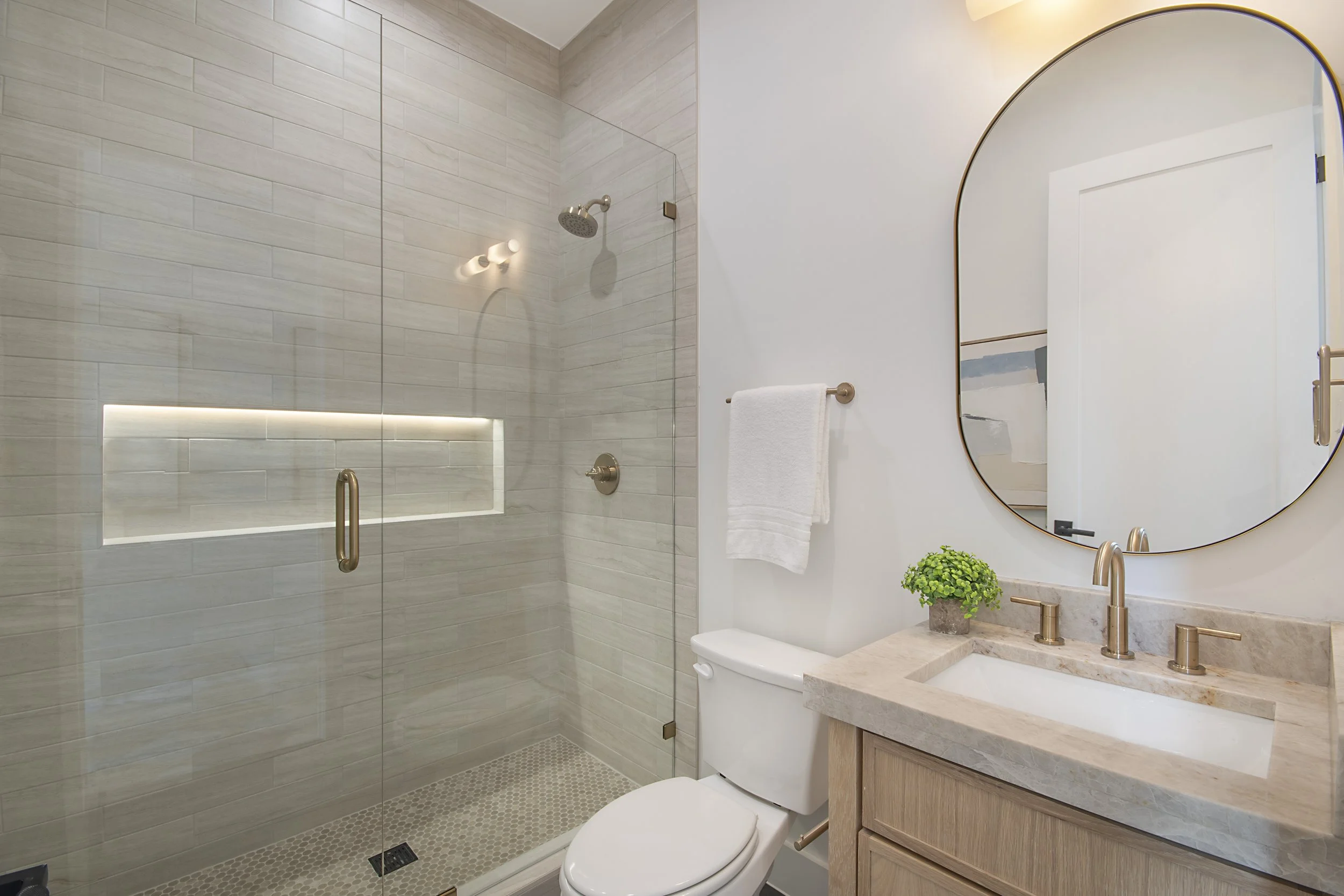 Modern bathroom with a glass-enclosed shower, beige tiles, a white toilet, a vanity with a marble countertop, a large round mirror, a small green plant, and a white towel hanging on a brass towel bar.