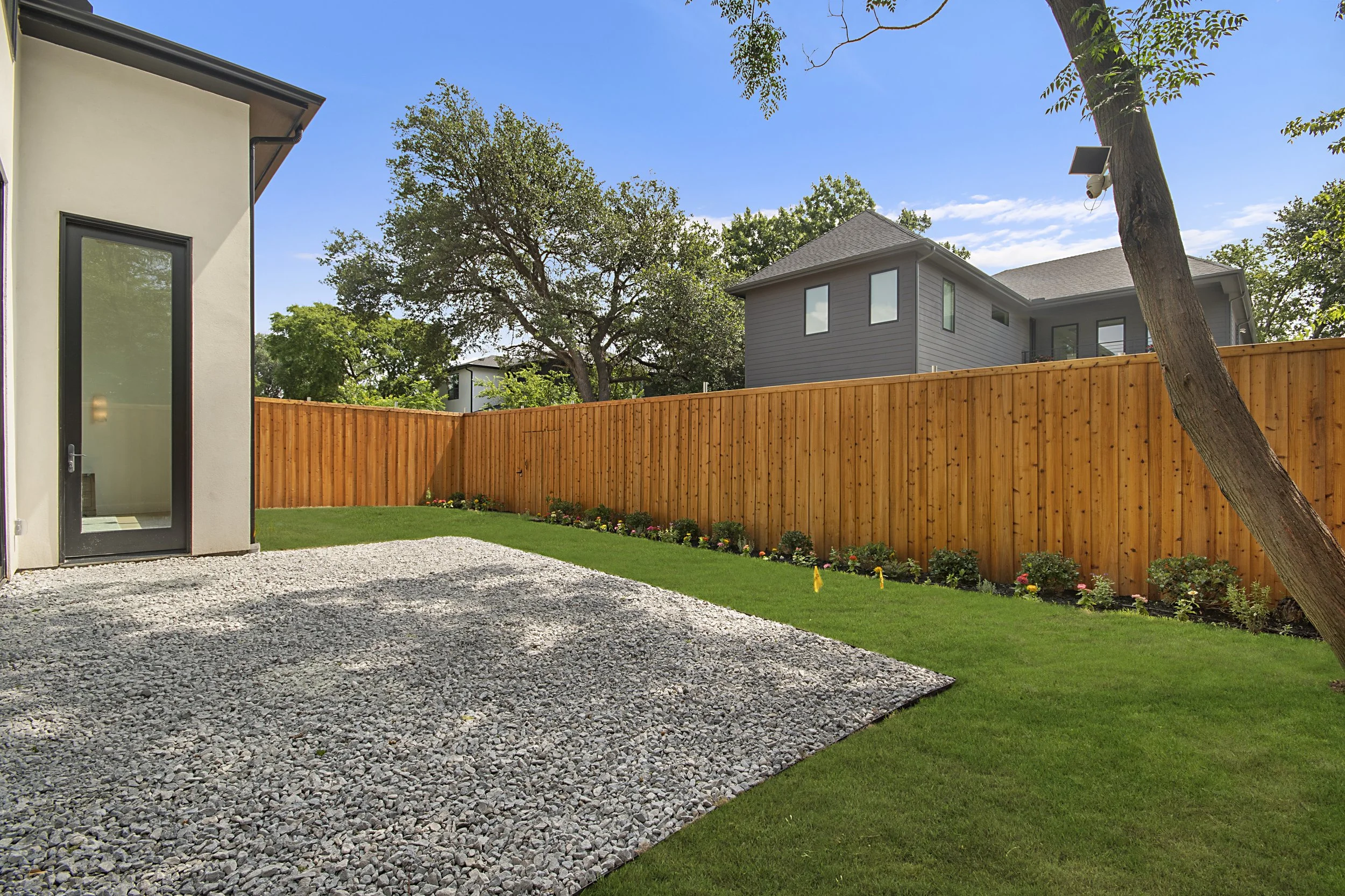 View of a backyard with a pebble patio, a green lawn, a tall wooden fence, mature trees, and a two-story house with gray siding and multiple windows, under a blue sky.