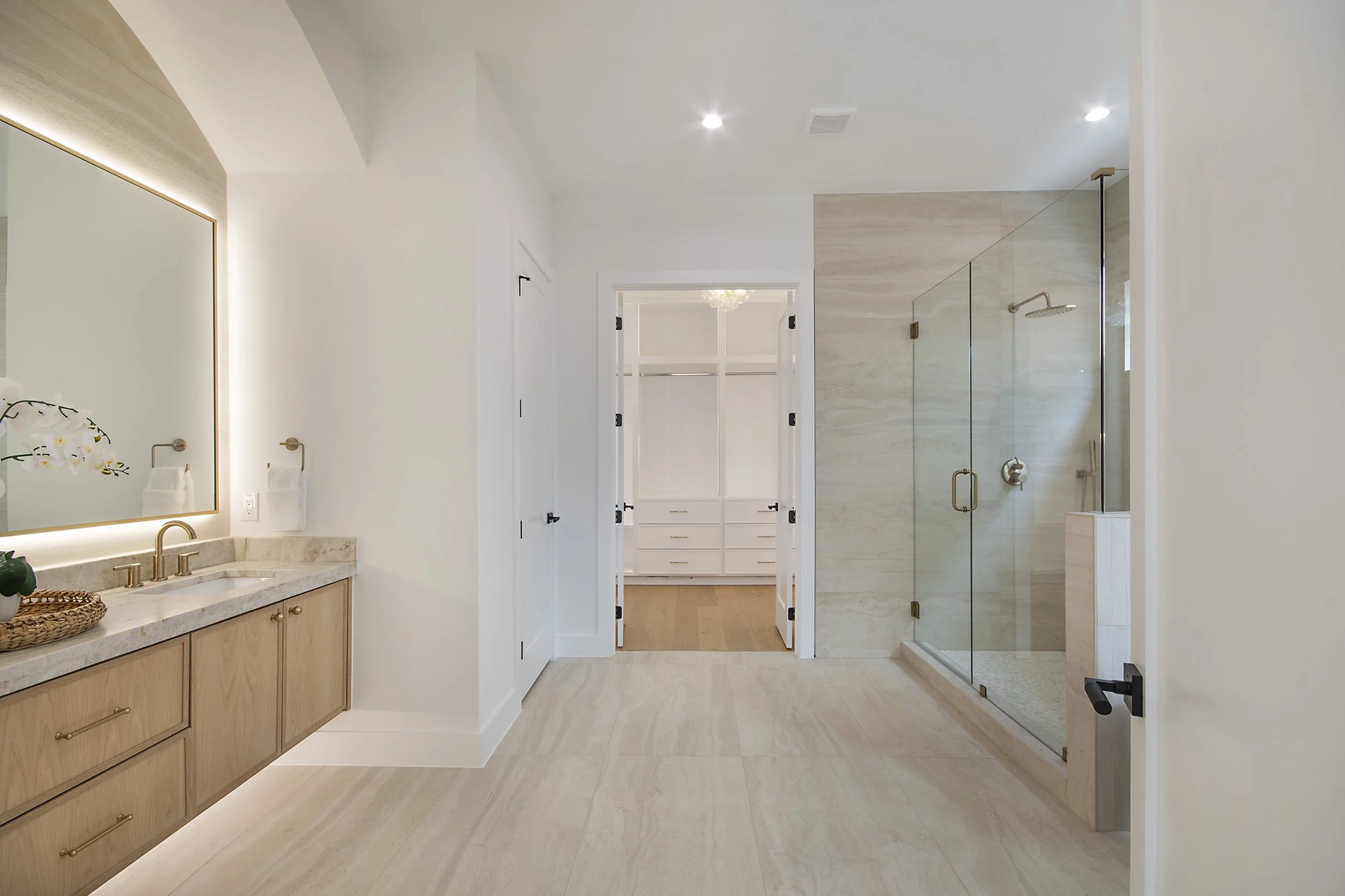 Modern bathroom with wooden vanity, large mirror, glass-enclosed shower, and separate toilet area visible through doorway.