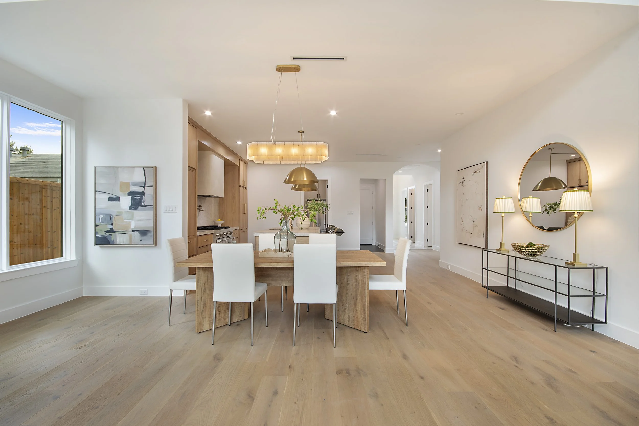 A modern open-concept kitchen and dining area with light wood flooring, white walls, a wooden dining table with white chairs, pendant lighting, and decorative mirrors and artwork.