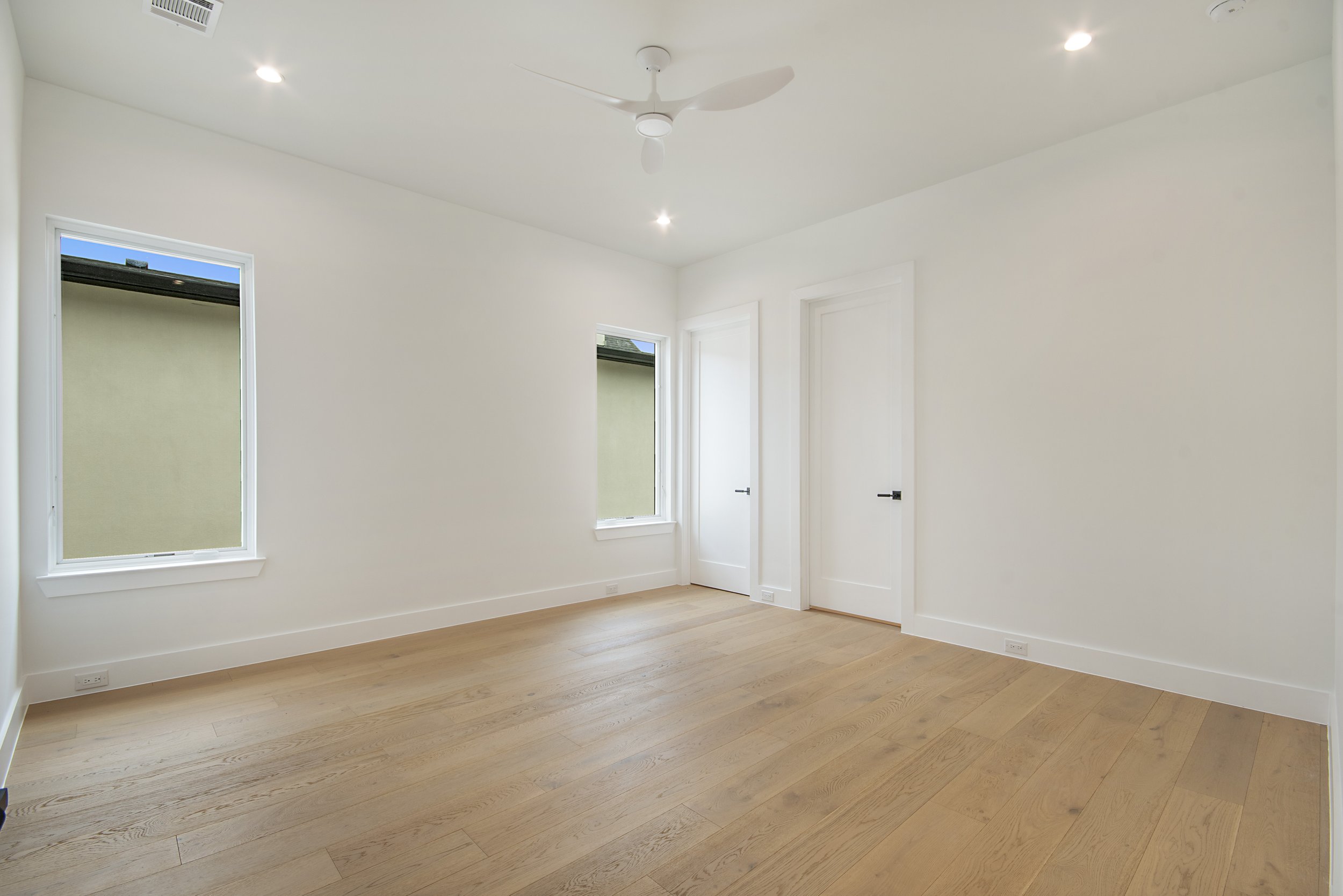 Empty room with white walls, hardwood floors, two windows, a door, a ceiling fan, and recessed lighting.
